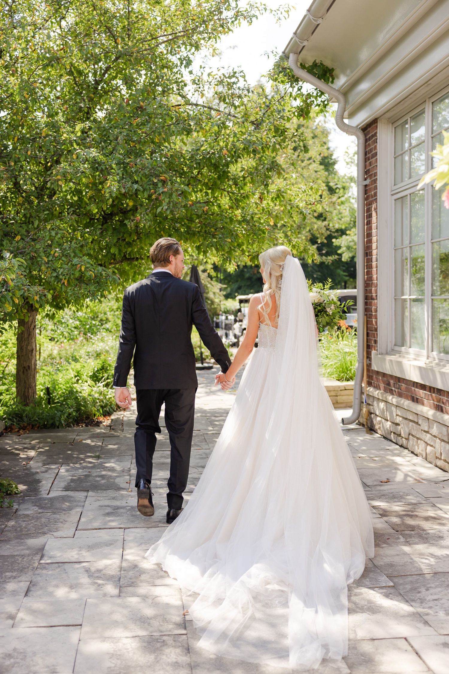 Newlyweds walking hand in hand along a stone path beside a brick building.