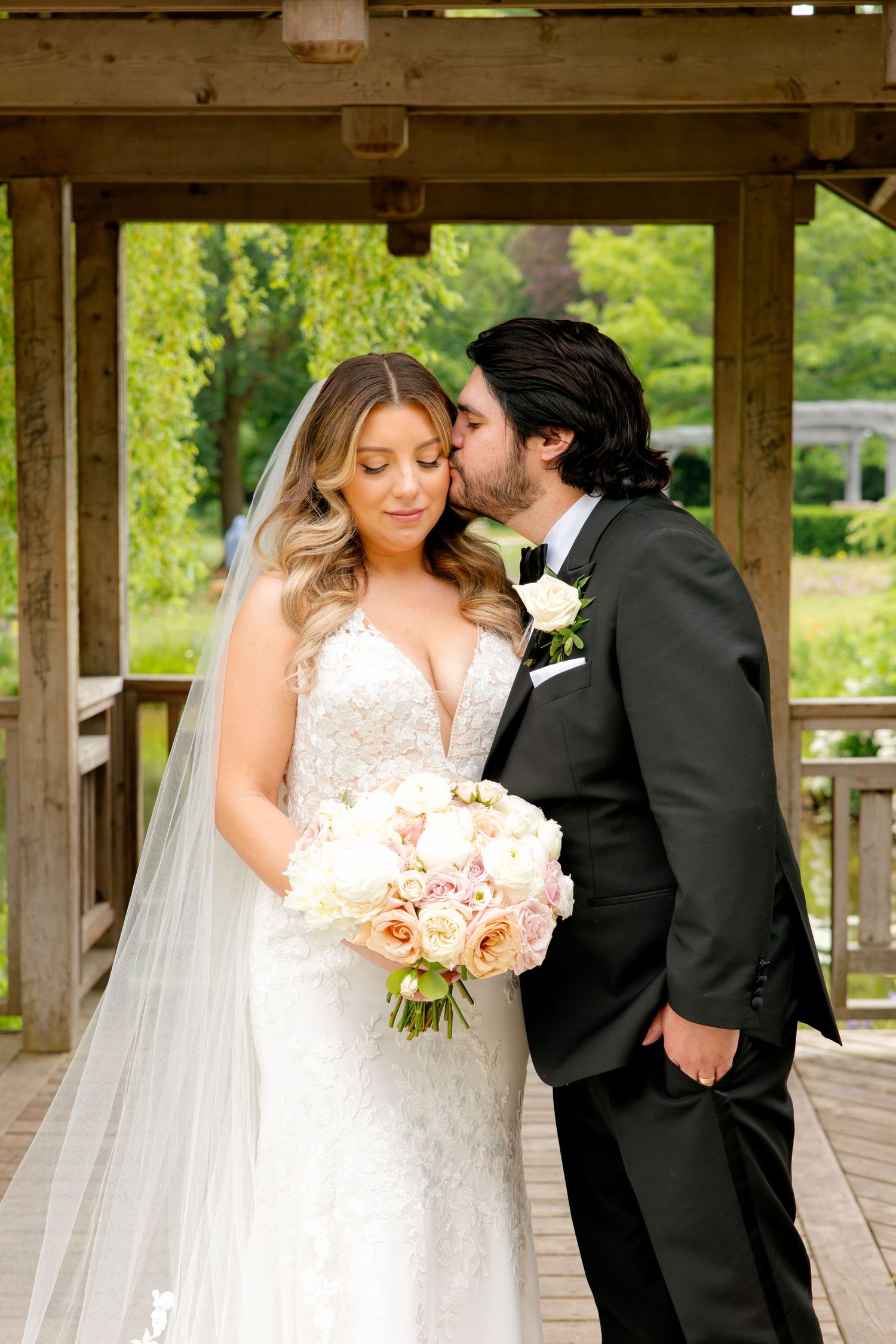Intimate wedding portrait of couple under wooden gazebo with bride holding pink bouquet.