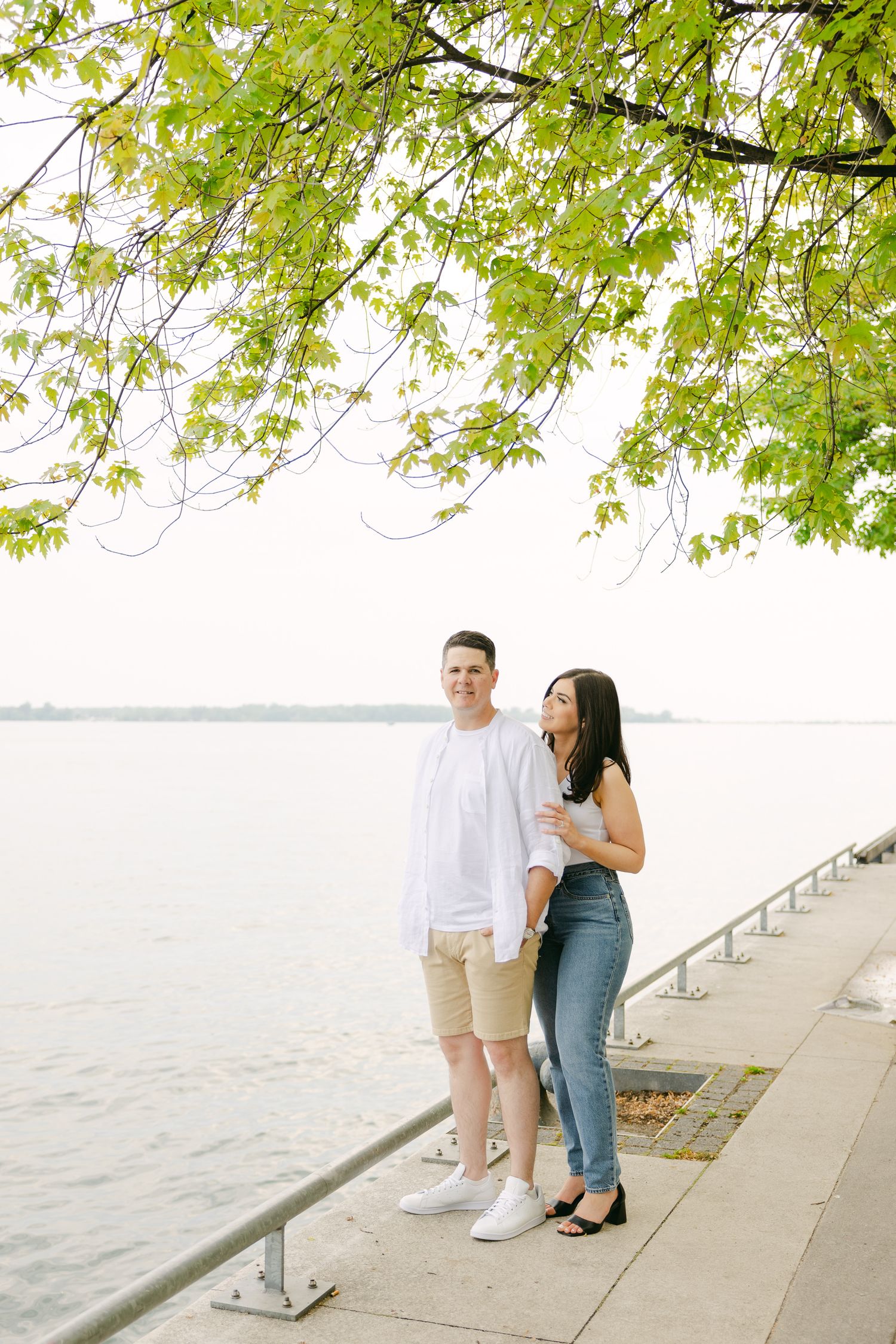 A couple stands together on a waterfront path under green spring leaves.