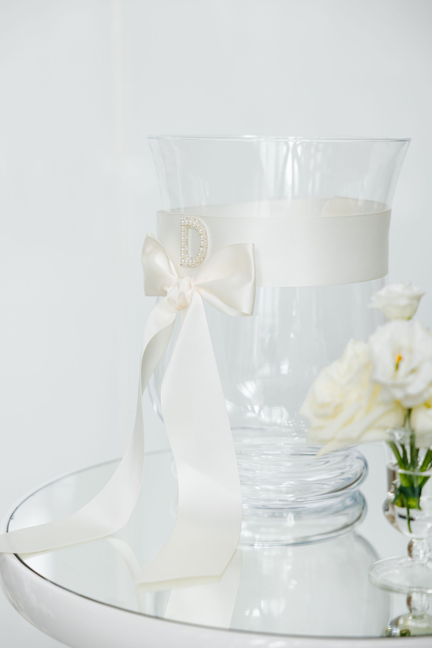 Glass vase decorated with white ribbon bow displayed on reflective surface with floral accents.