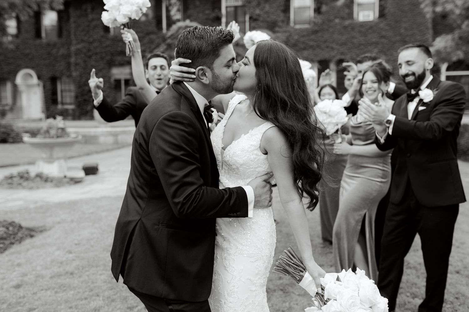 Black and white photo of newlyweds kissing while wedding party celebrates in background.