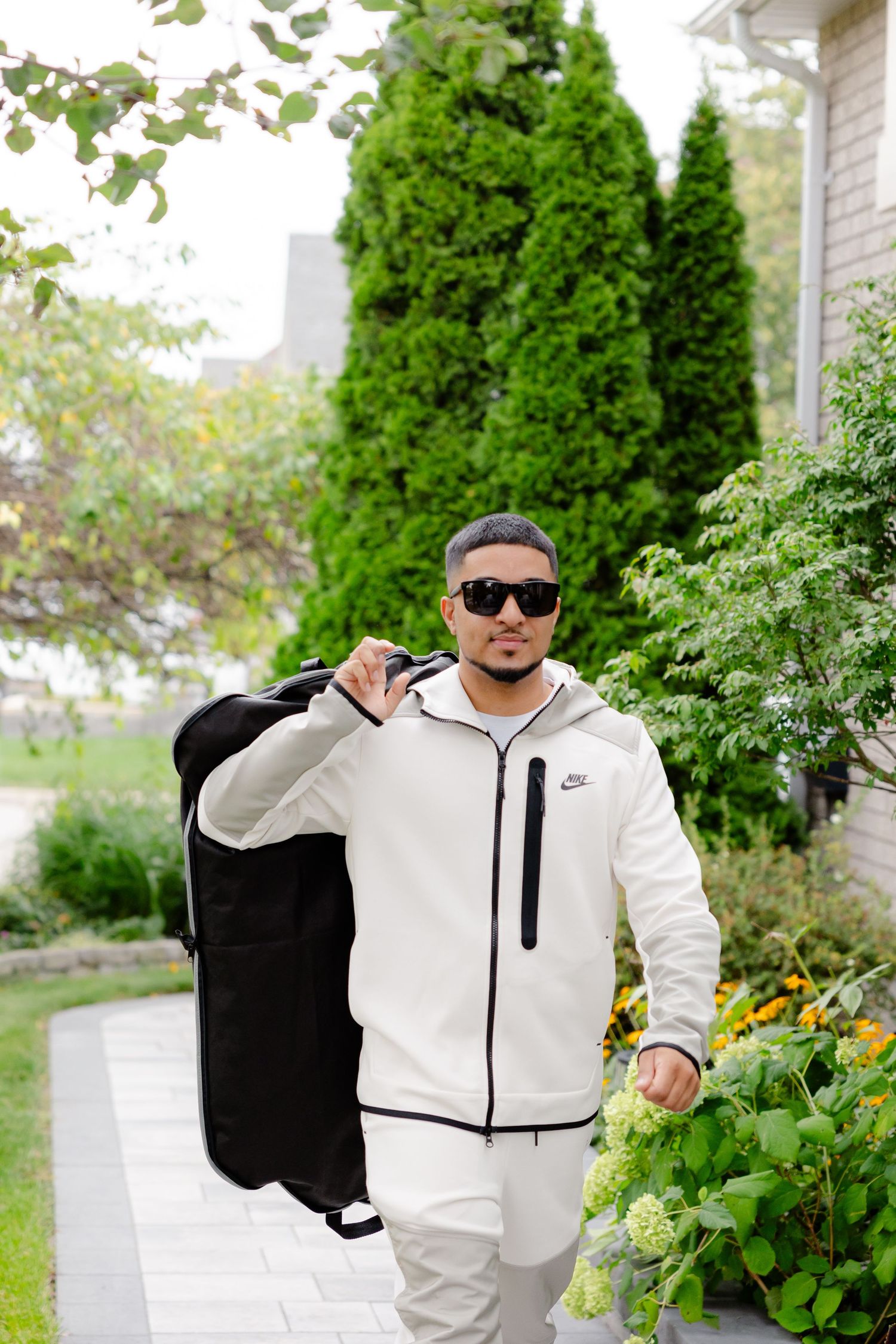 A person in white athletic wear carries a black garment bag outside a house with evergreen trees.