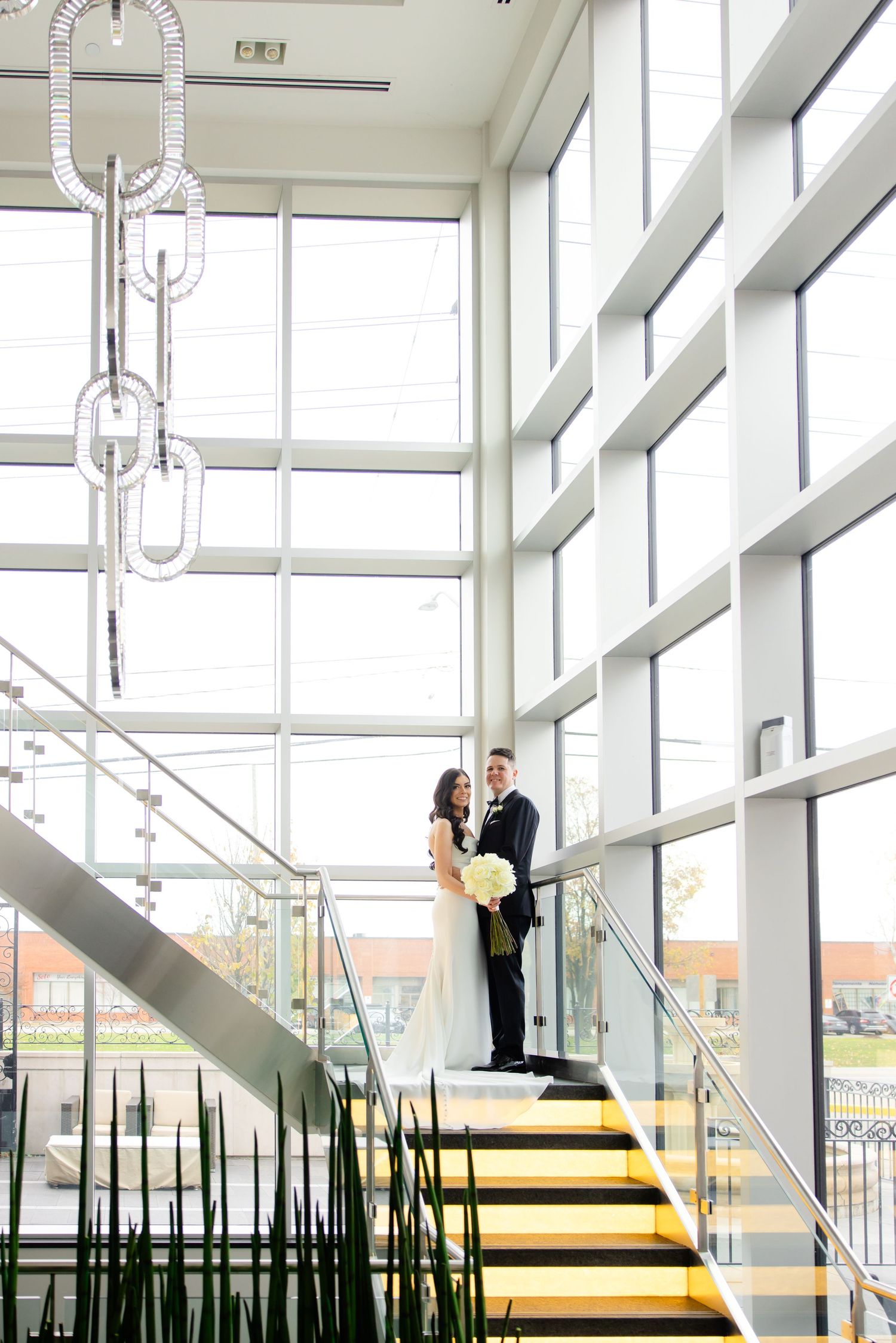 A couple stands elegantly on yellow-lit stairs in a modern building with floor-to-ceiling windows and hanging lights.