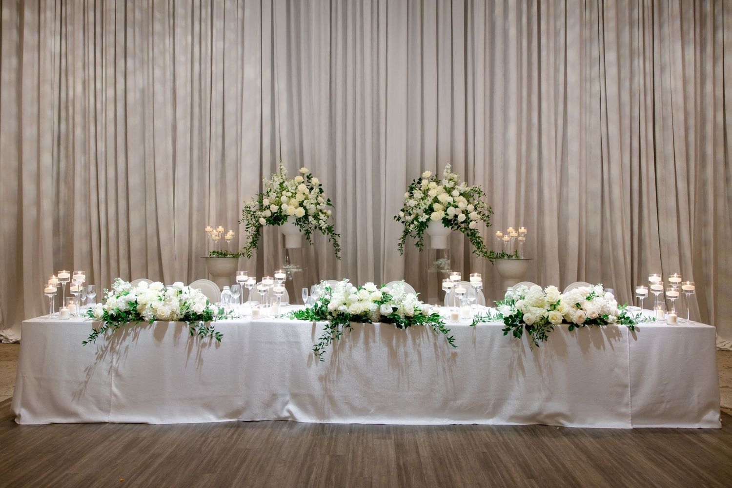 White wedding head table display with elevated floral arrangements and candlelight against draped backdrop.