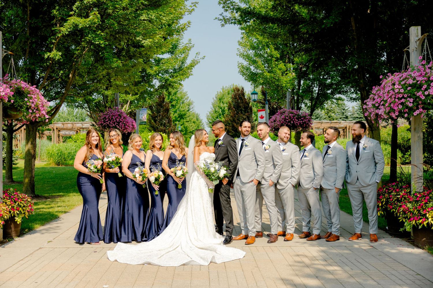 Wedding party in navy dresses and gray suits poses on garden path with flowers.