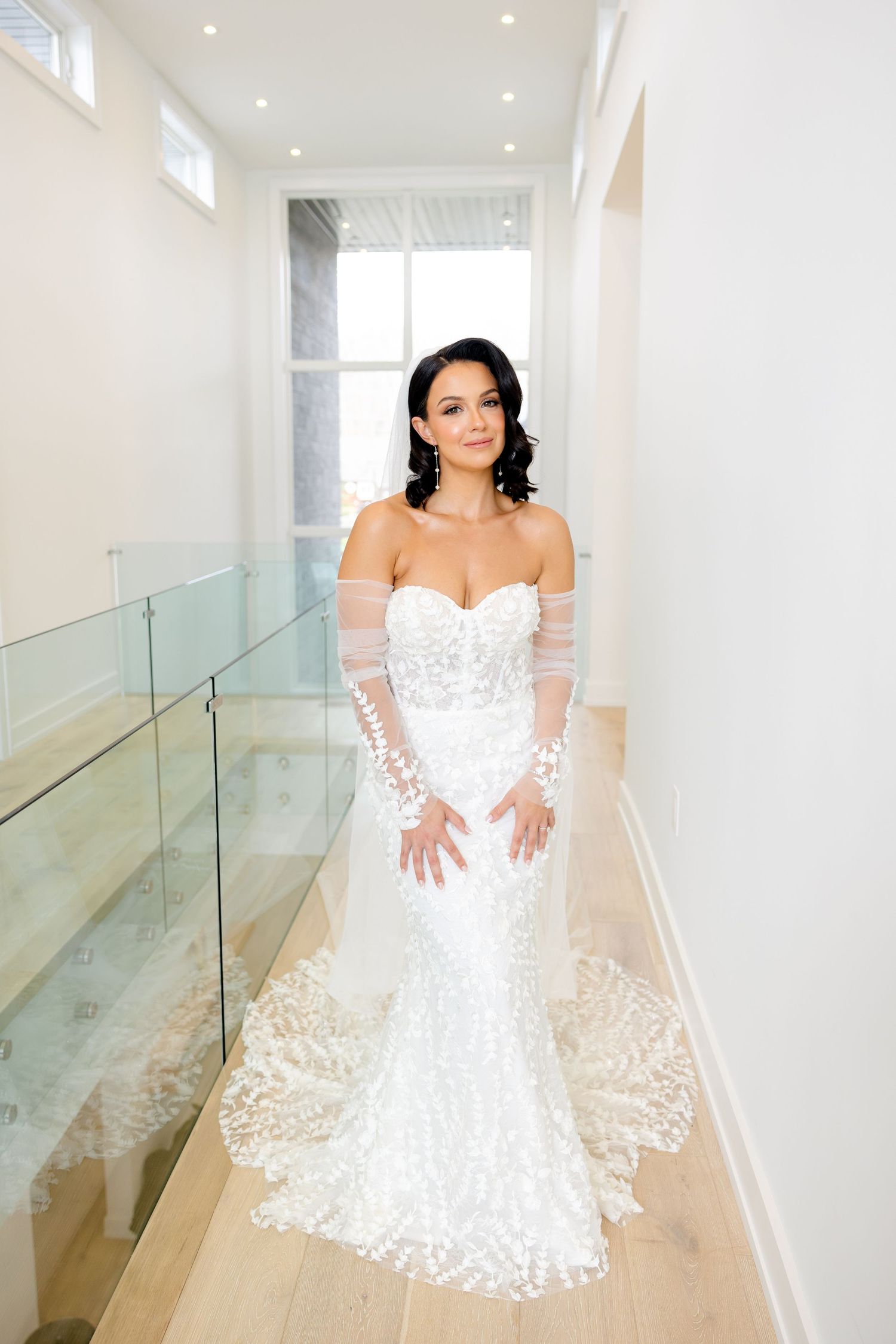 Bride in form-fitting lace gown poses in modern hallway with glass railing and natural light.