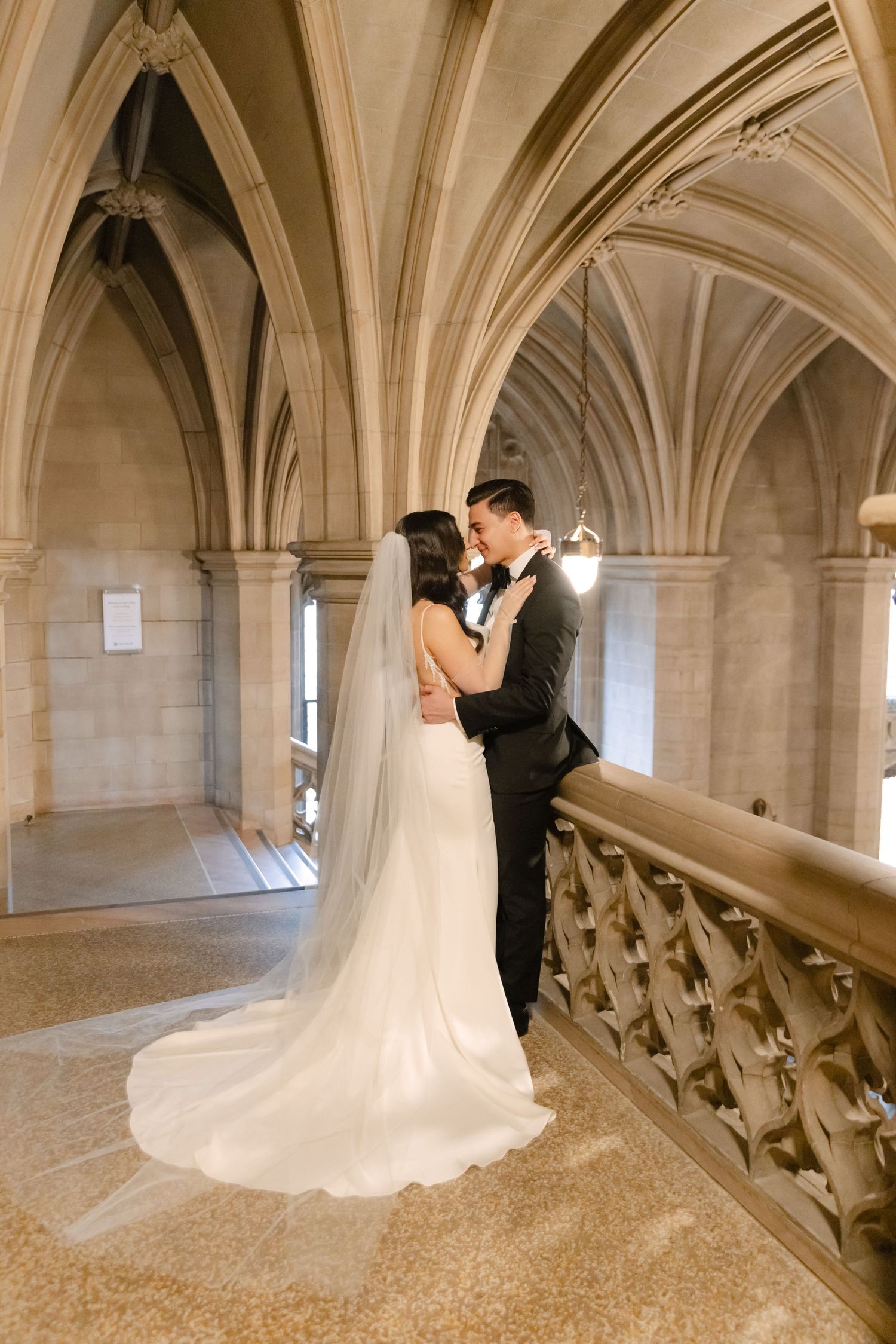 Elegant wedding couple embraces on historic stone balcony under vaulted ceiling.