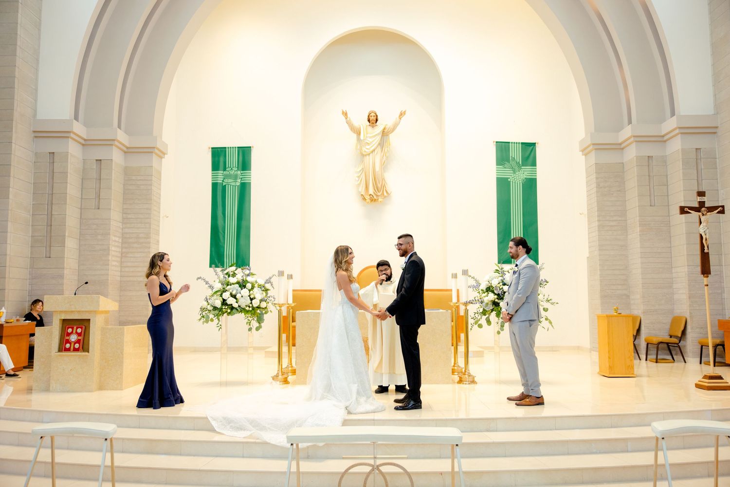 Wedding ceremony taking place in a beautiful Catholic church with green accents and white altar decorations.