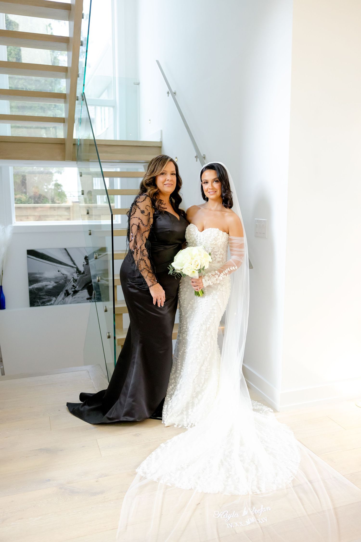 Two women pose on a modern staircase, one in a black gown and one in a white wedding dress with bouquet.