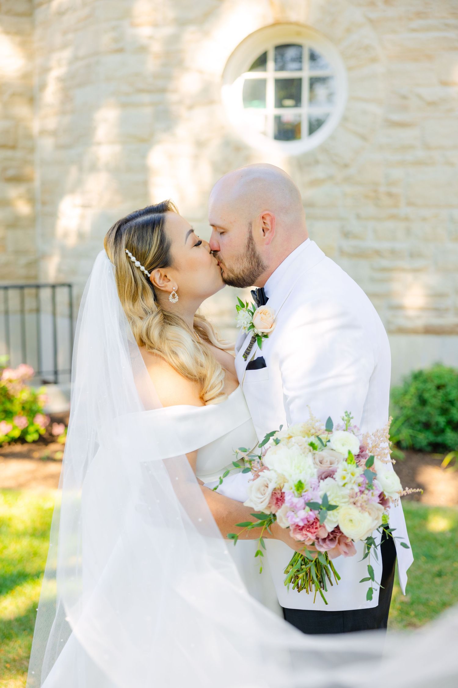 A romantic wedding kiss captured in sunlight against a stone building with a round window.