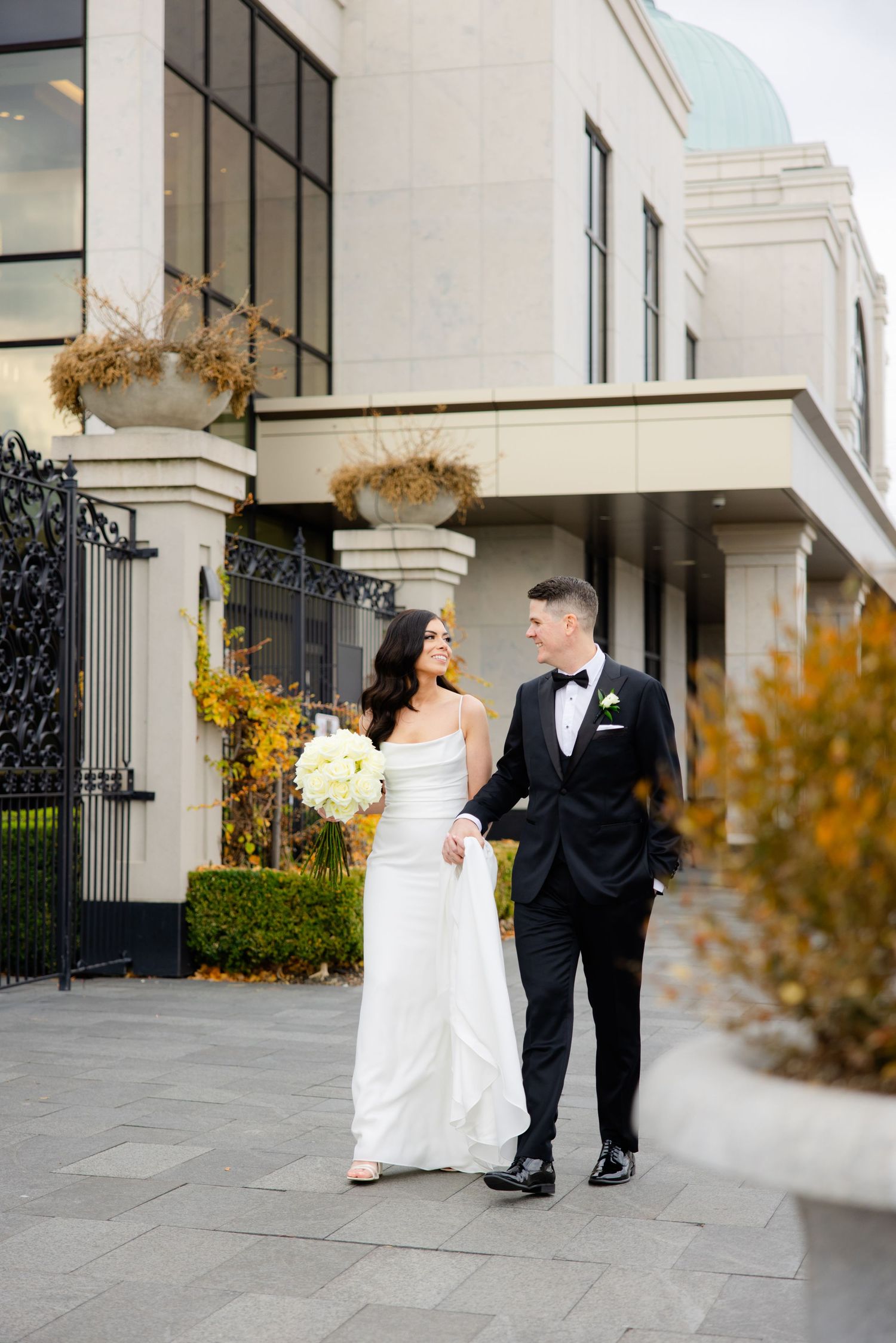 Newlyweds share a moment outside modern building with autumn decorative planters and iron gates.