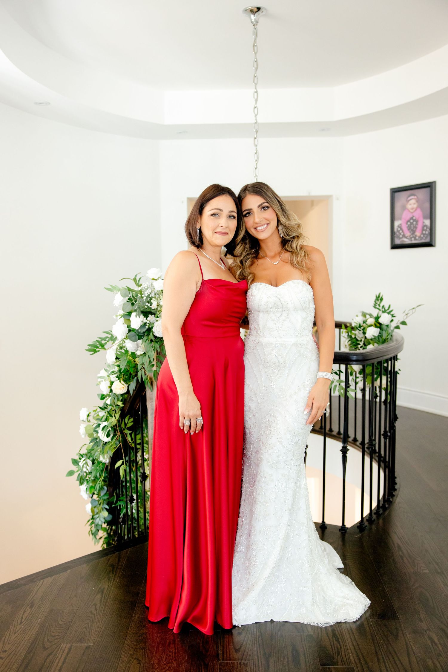 Wedding photo featuring a bride in white lace gown and bridesmaid in red floor-length dress with floral decor.