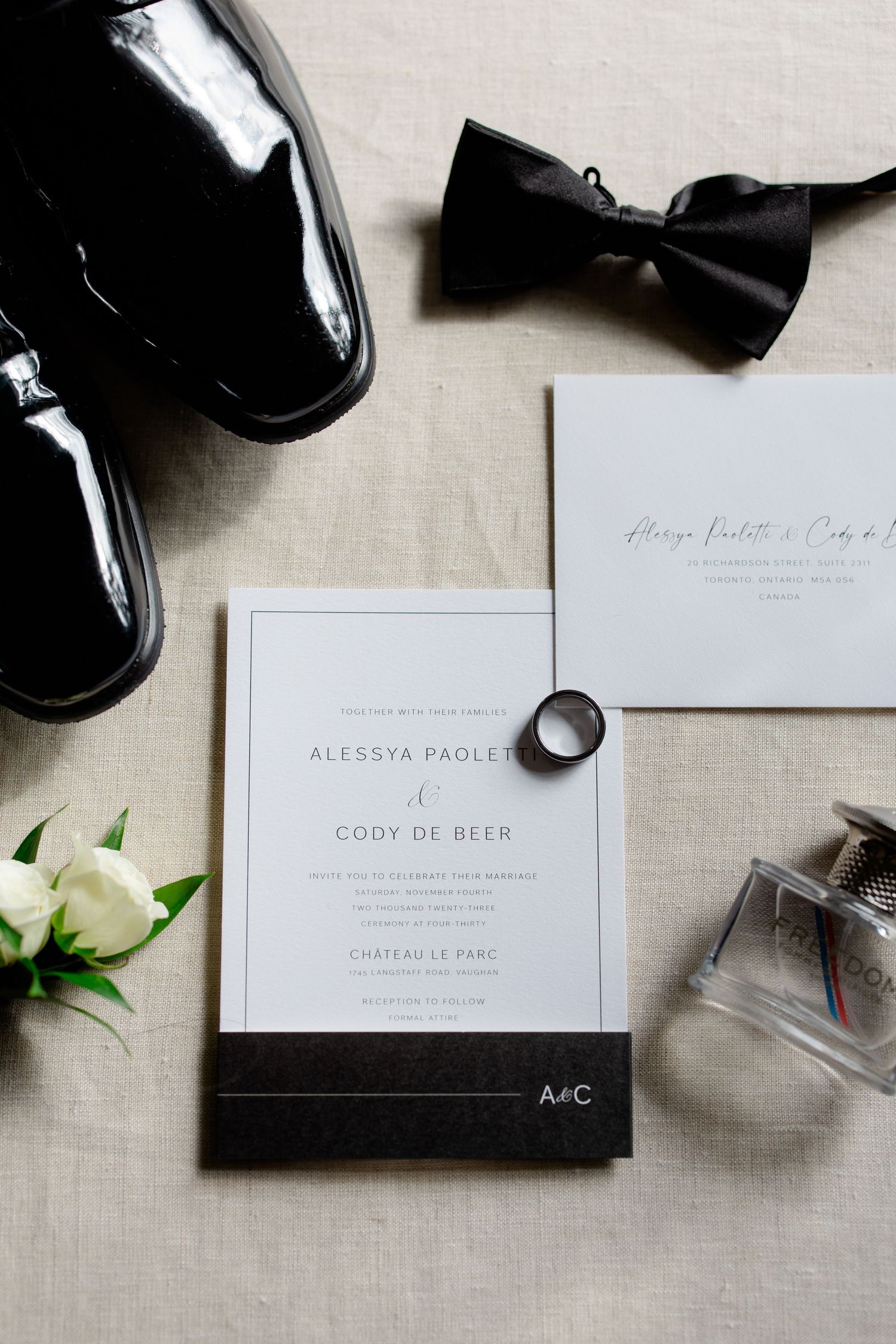 Wedding details including patent leather shoes, black bow tie, invitation and floral accent on linen background.