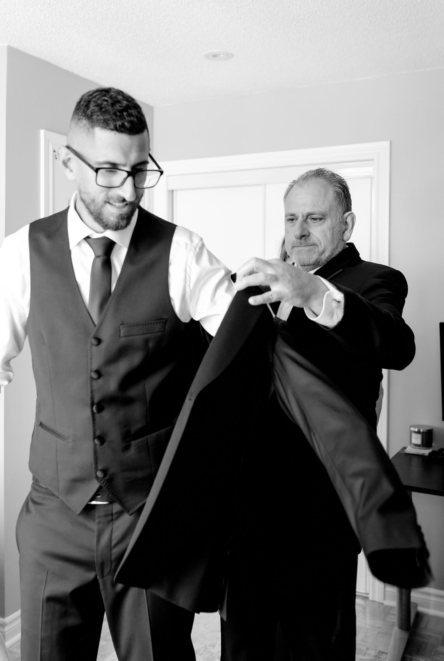 Two people in formal wear help adjust a suit jacket during preparations.