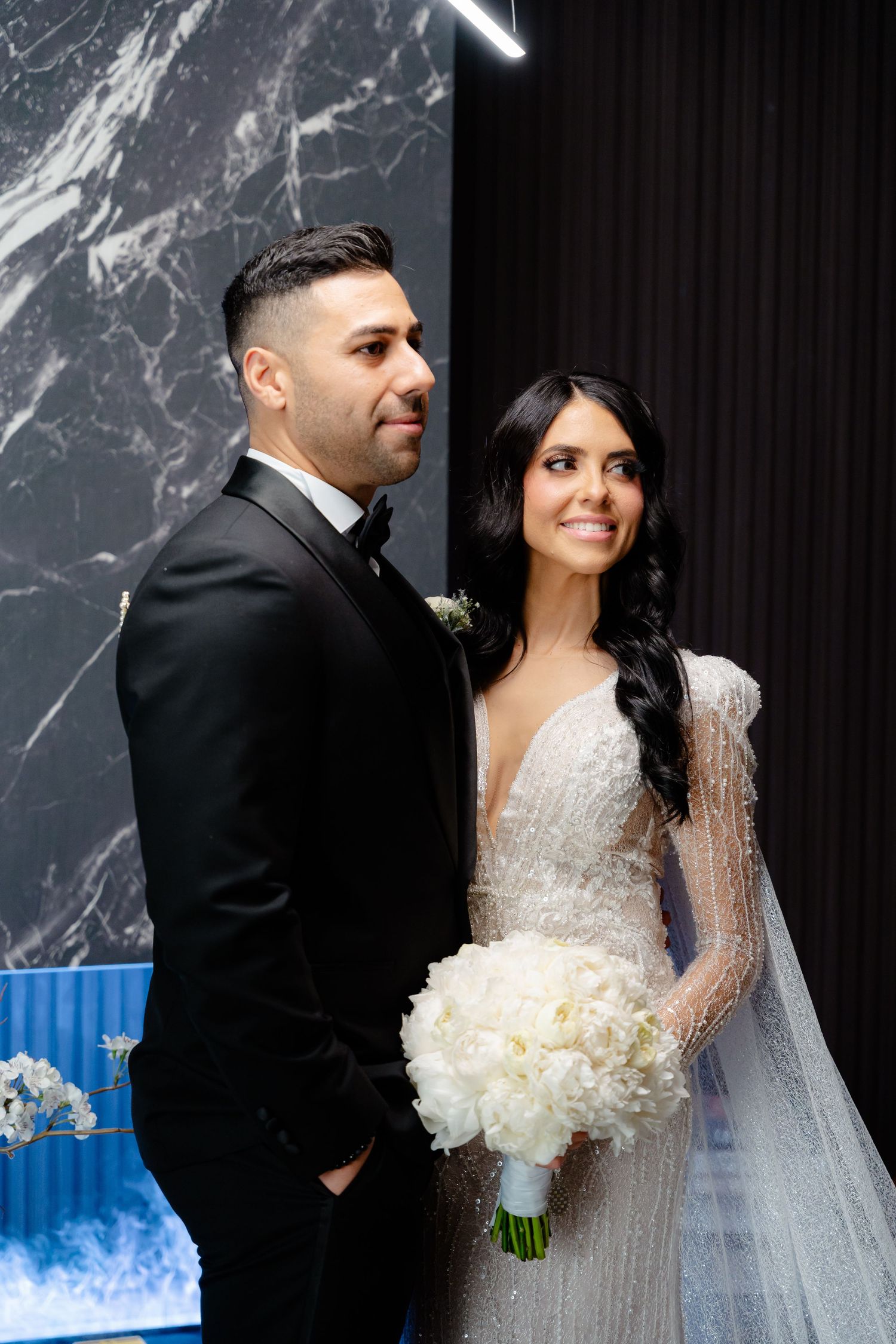 A wedding couple poses against a marble wall backdrop, the bride holding a white peony bouquet.