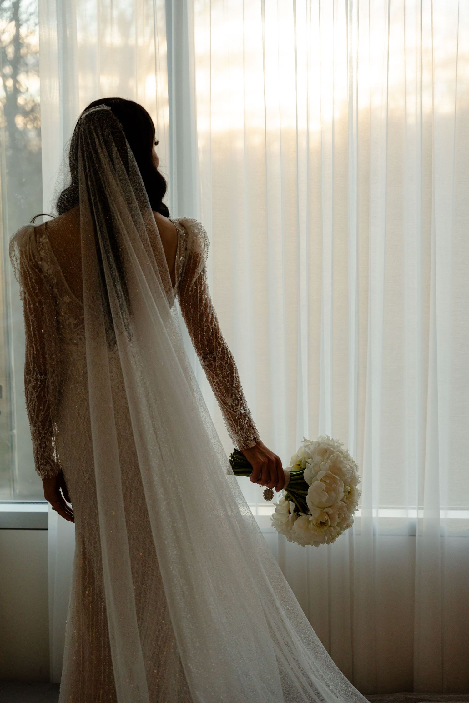 Silhouette of bride holding white bouquet in lace gown with long veil against window.