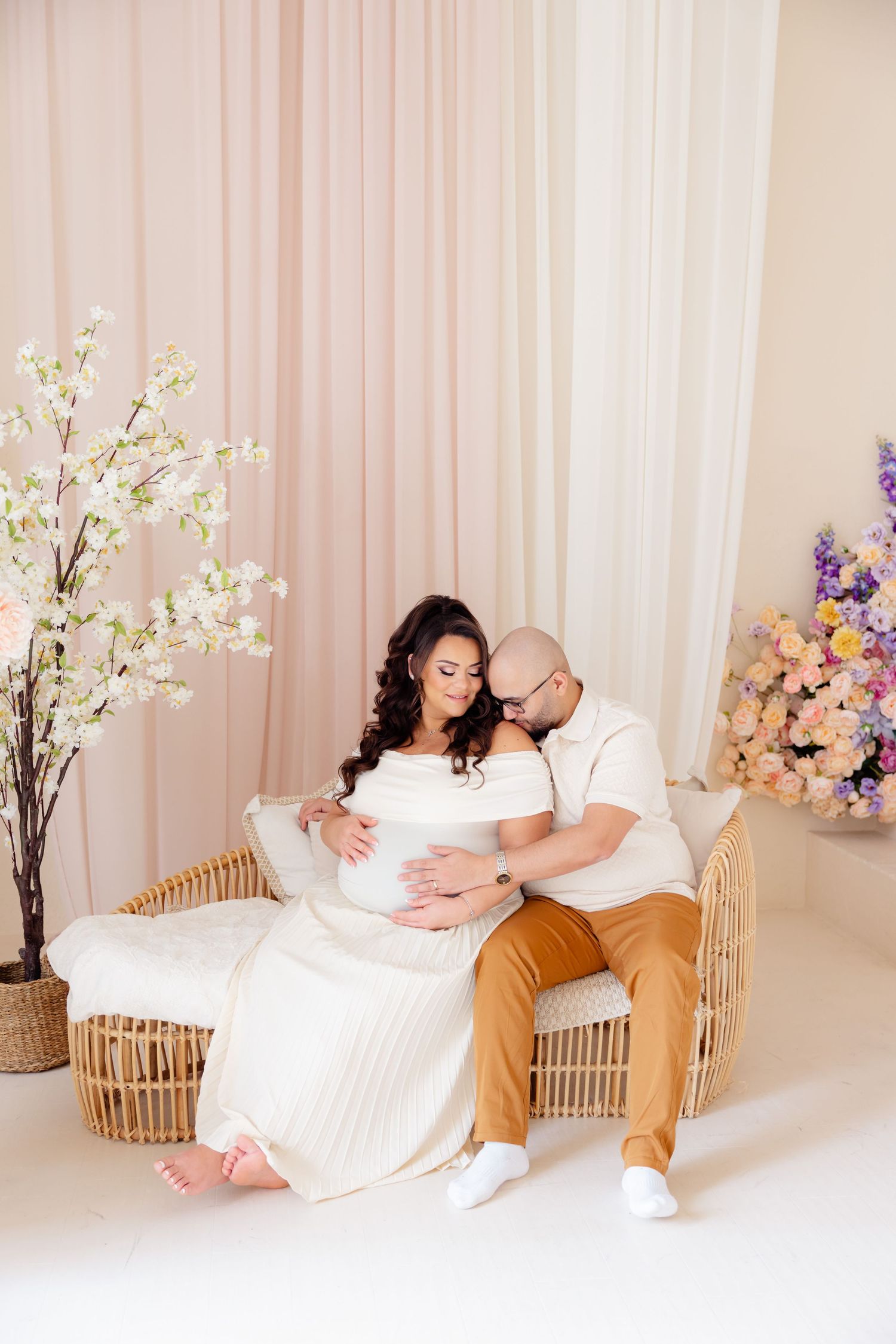 Romantic maternity photo with couple sitting on rattan bench surrounded by spring floral arrangements in soft pink studio setting.