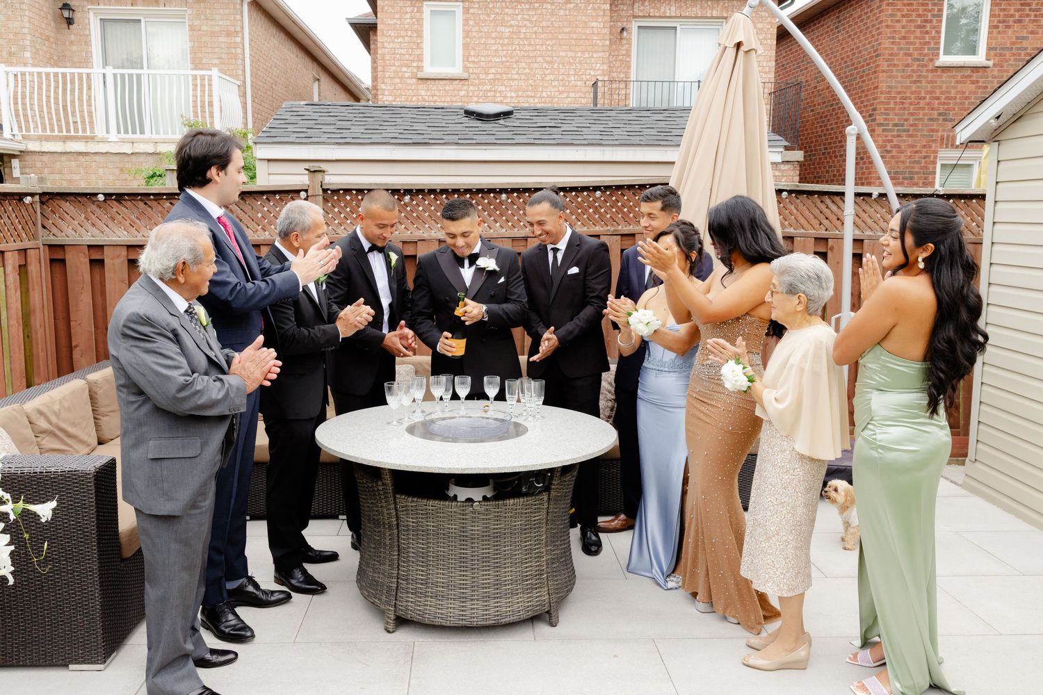 A celebration scene on an outdoor patio with people in formal wear gathered around a circular fire table.