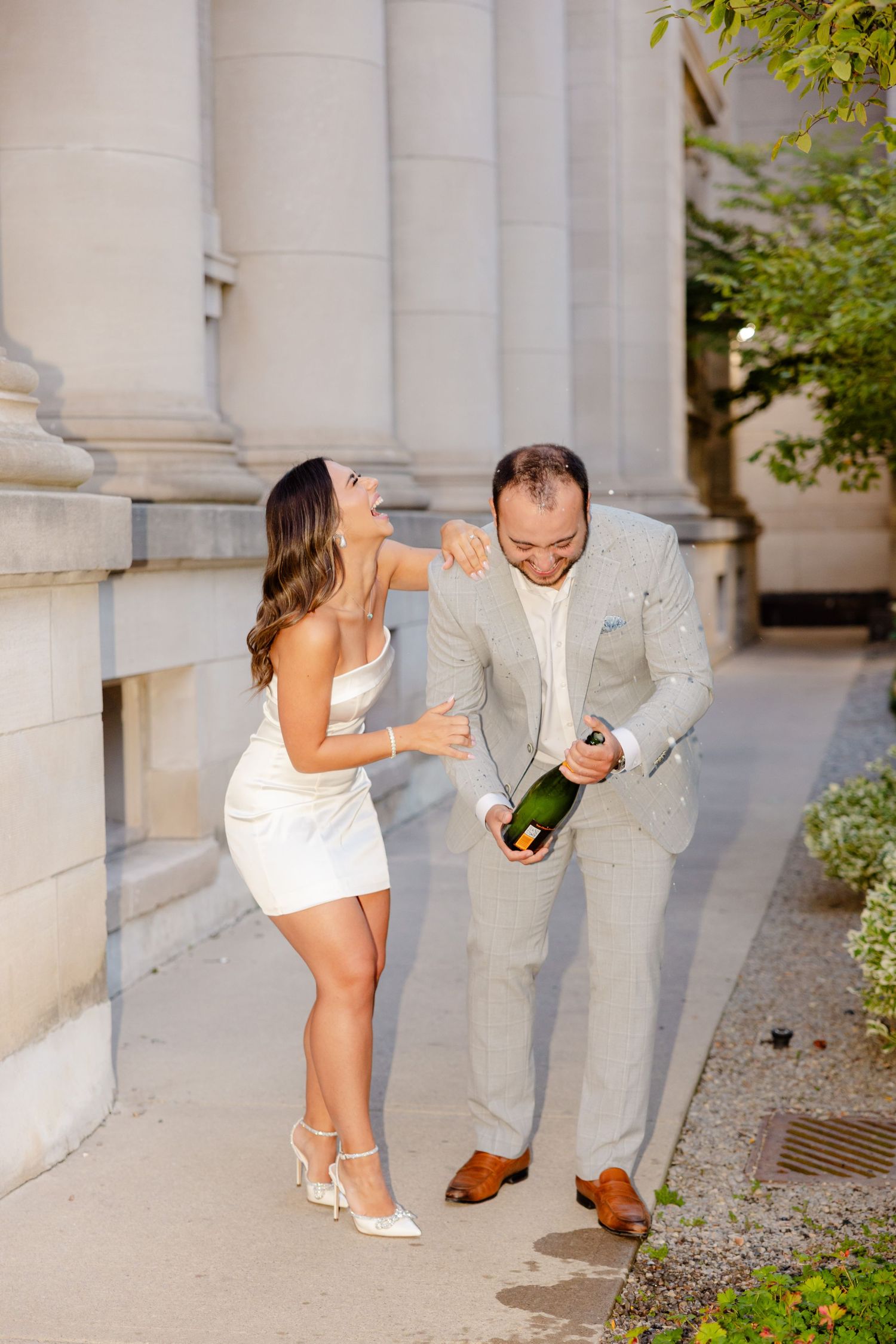 A couple laughs together while celebrating with champagne beside stone pillars.