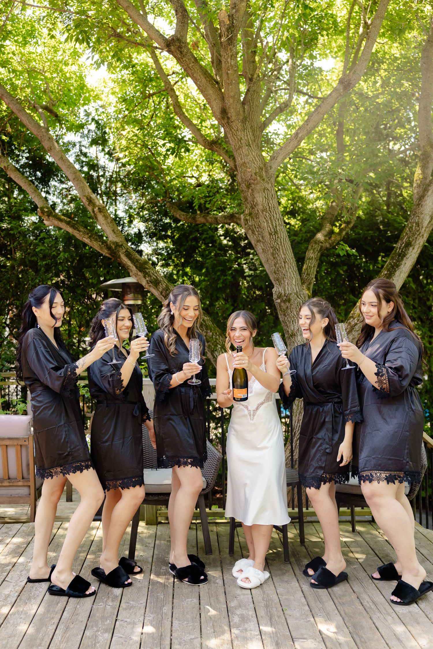 A bridal party in matching black robes laughing together on a wooden deck under sunlit trees.