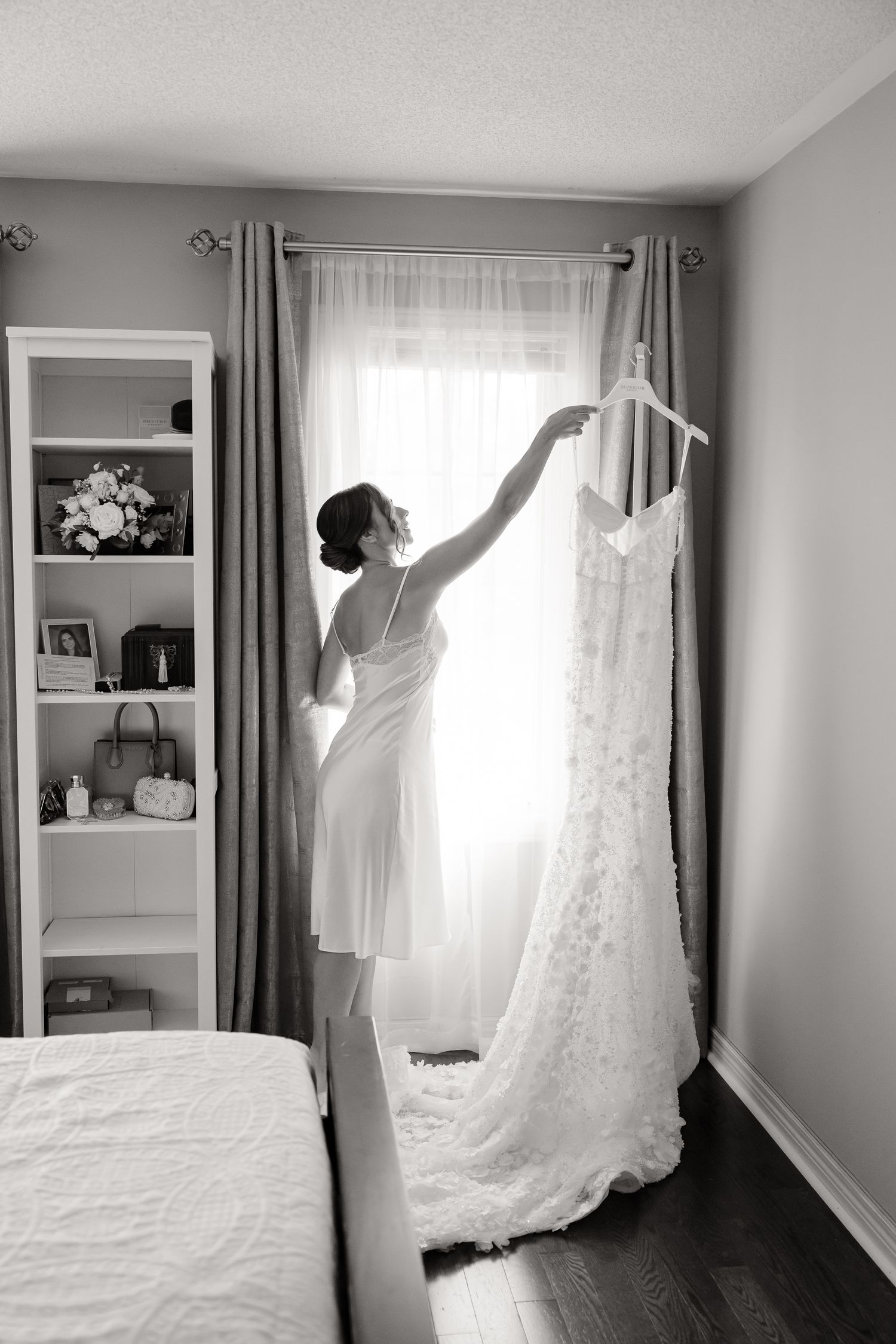 A white lace wedding gown hangs elegantly by a window as someone reaches up to touch it in a black and white photo.