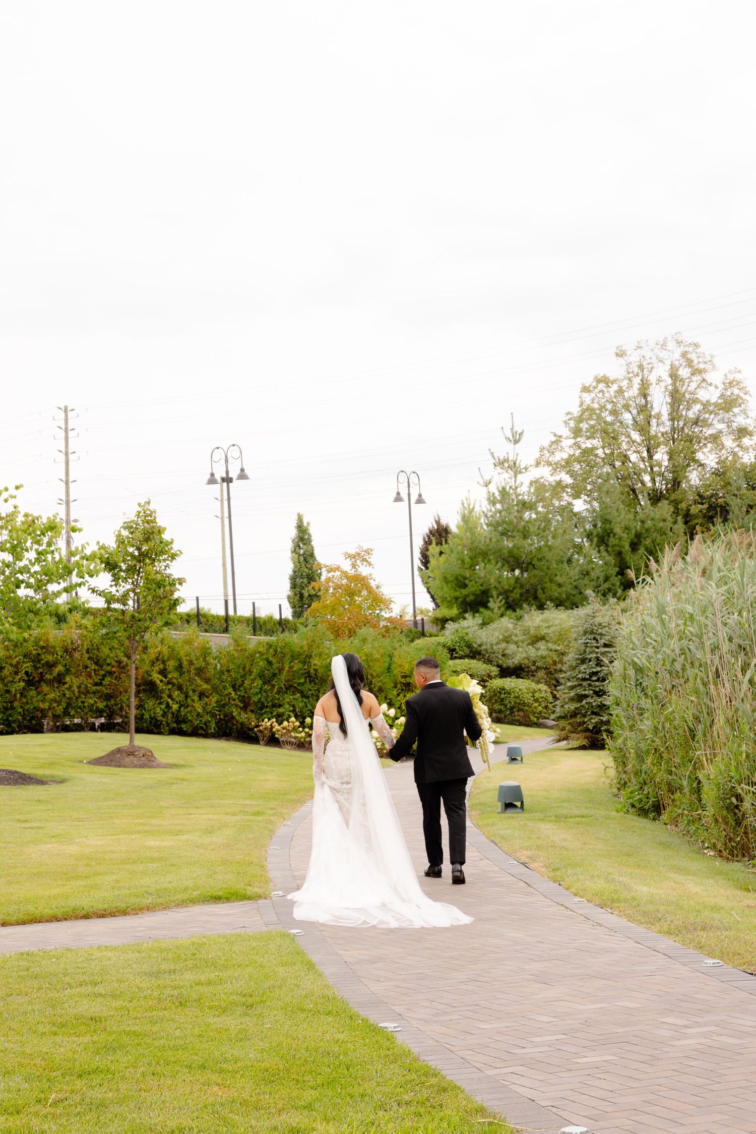 Couple walking together on a winding brick path through a lush garden setting with trees and greenery around them.