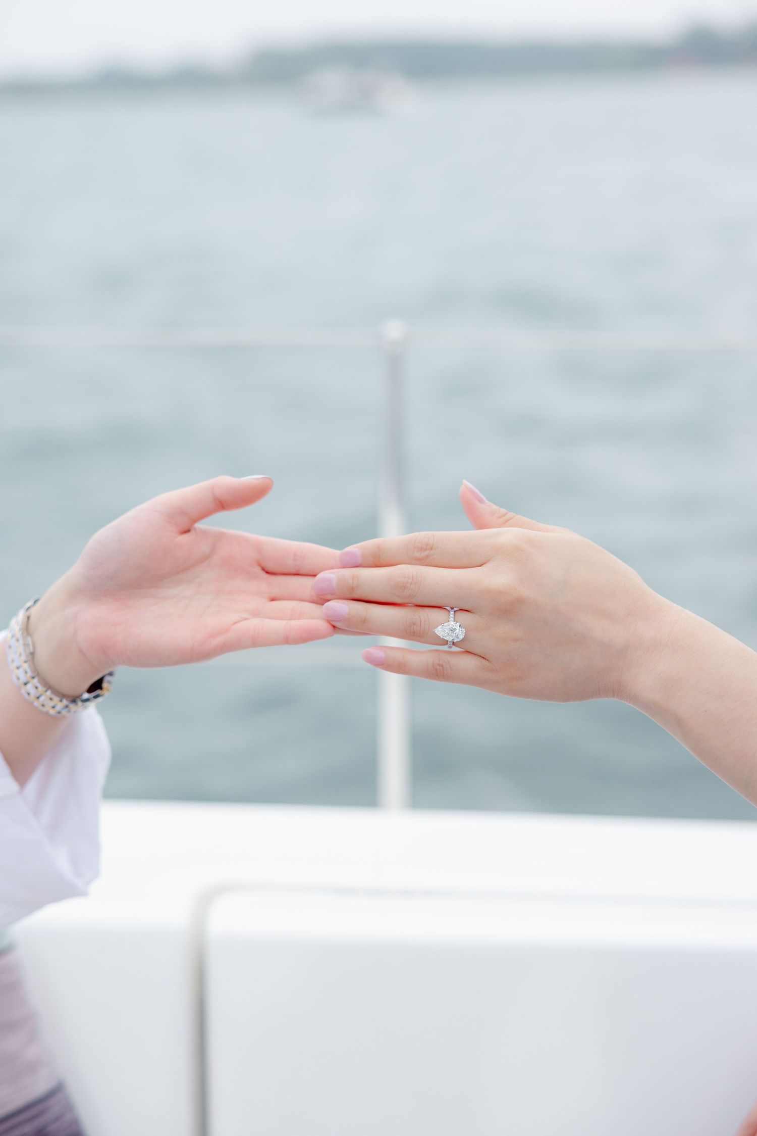 Close-up of two hands touching, with one wearing an engagement ring, against a blurry ocean background.