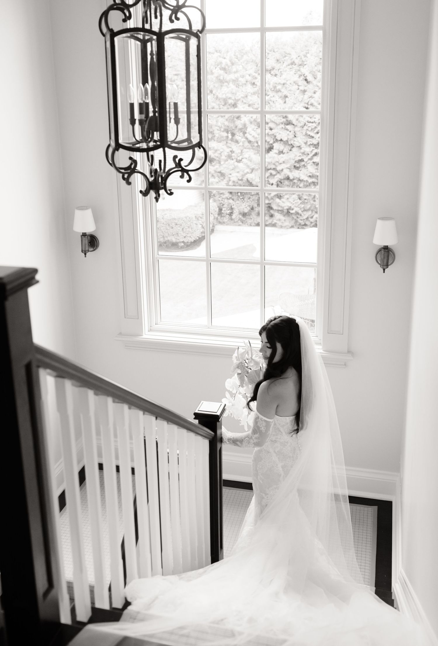 Black and white photograph showing a bride in a flowing white gown and veil standing by a grand window with an ornate chandelier.