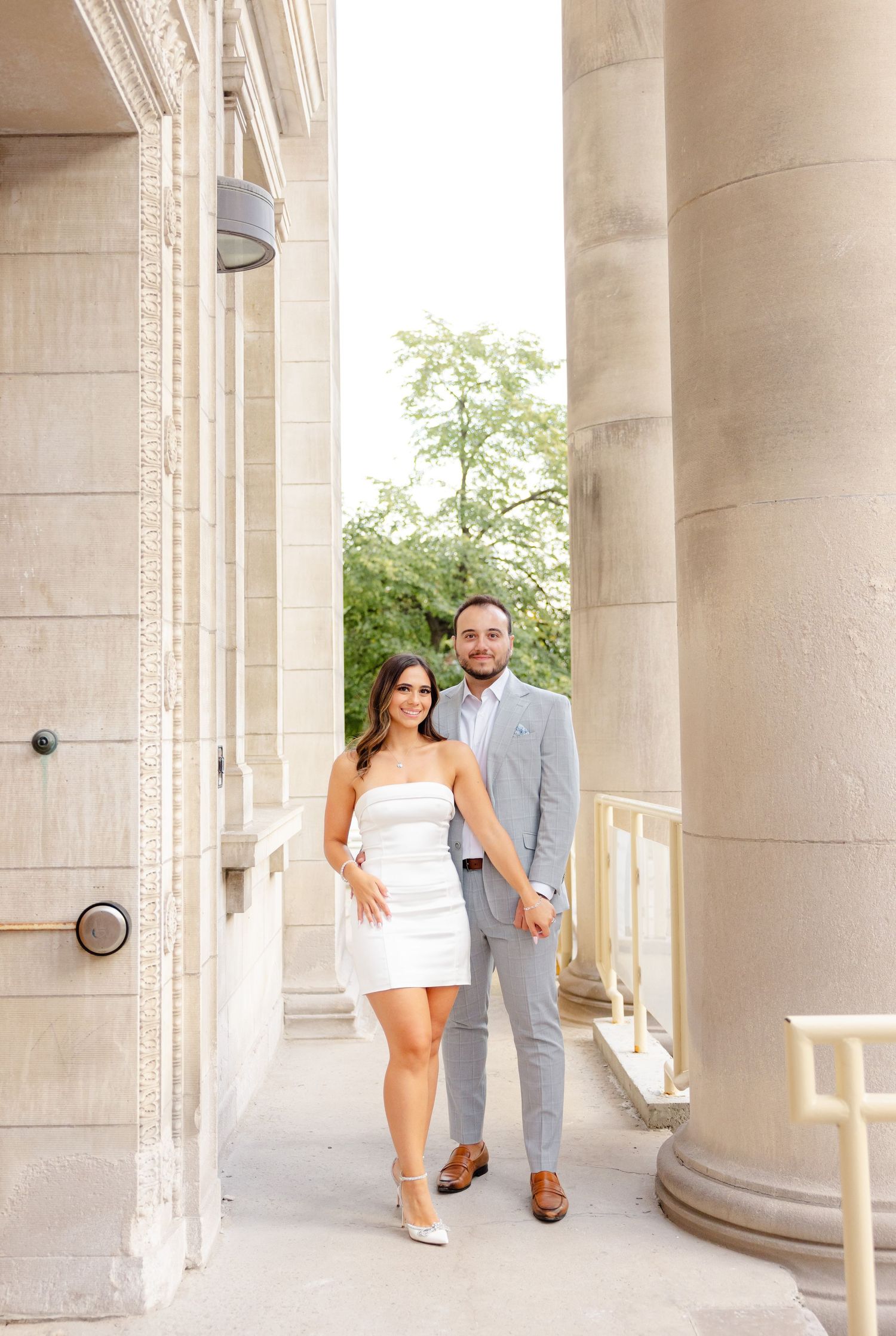 Couple in formal attire posing between classical stone columns with natural lighting.