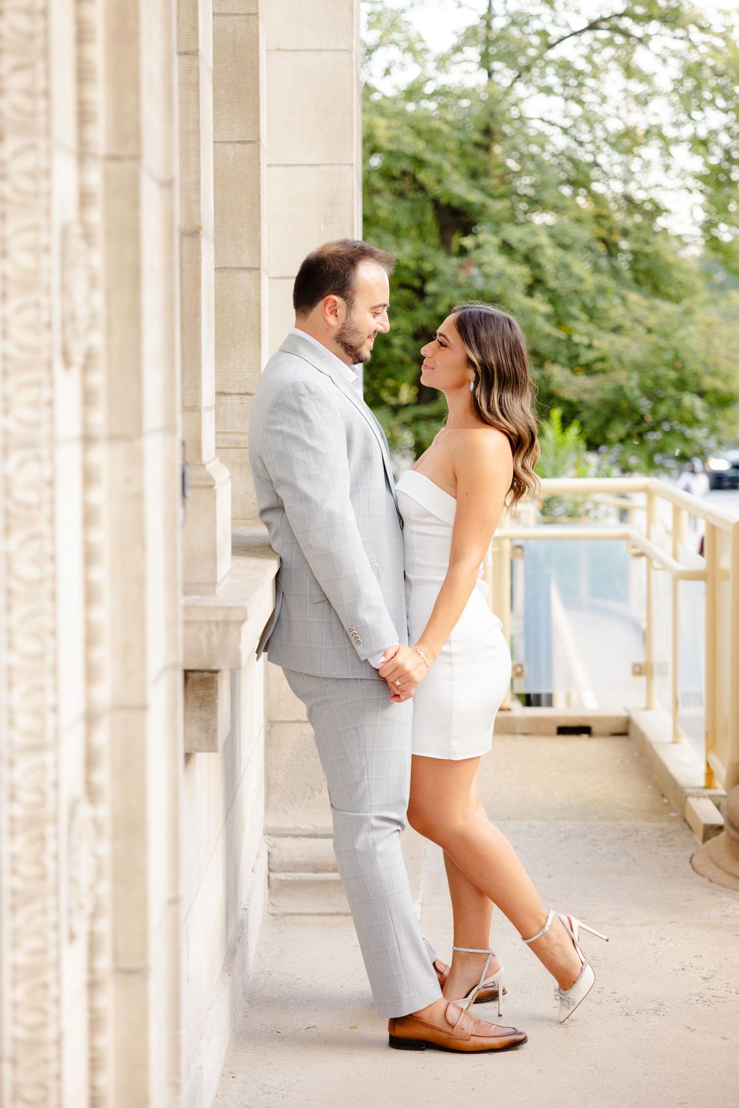 A stylish couple shares an intimate moment on a columned porch, her in white and him in light gray.