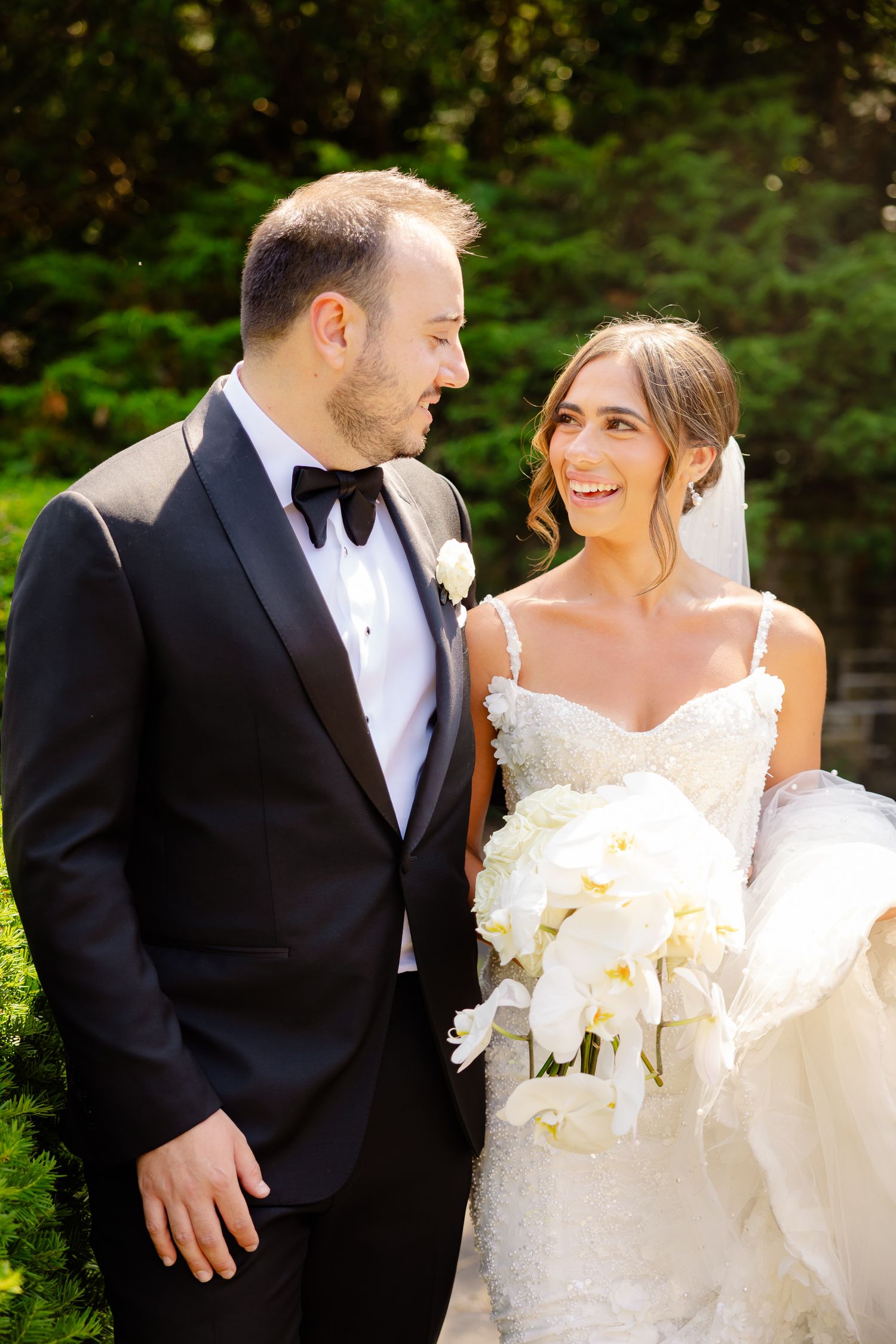 The bride and groom share joyful laughter during their wedding photo session in a verdant garden setting.