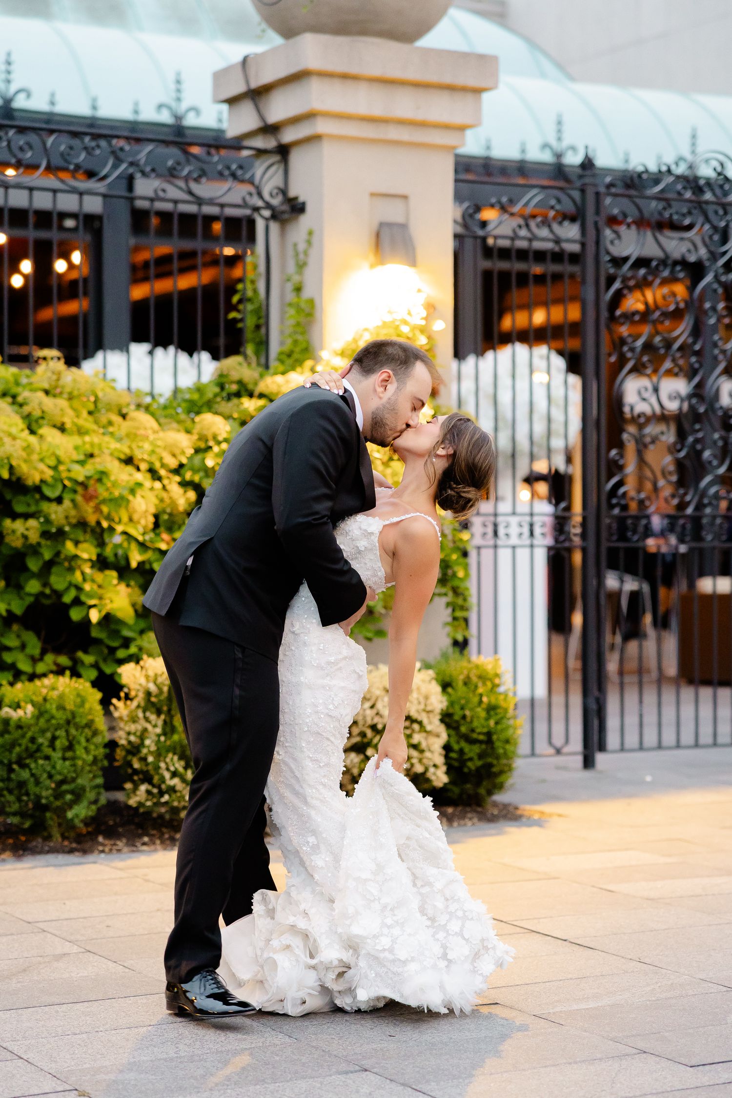 A romantic wedding couple share a kiss in front of an ornate iron gate with yellow flowers and stone pillars.
