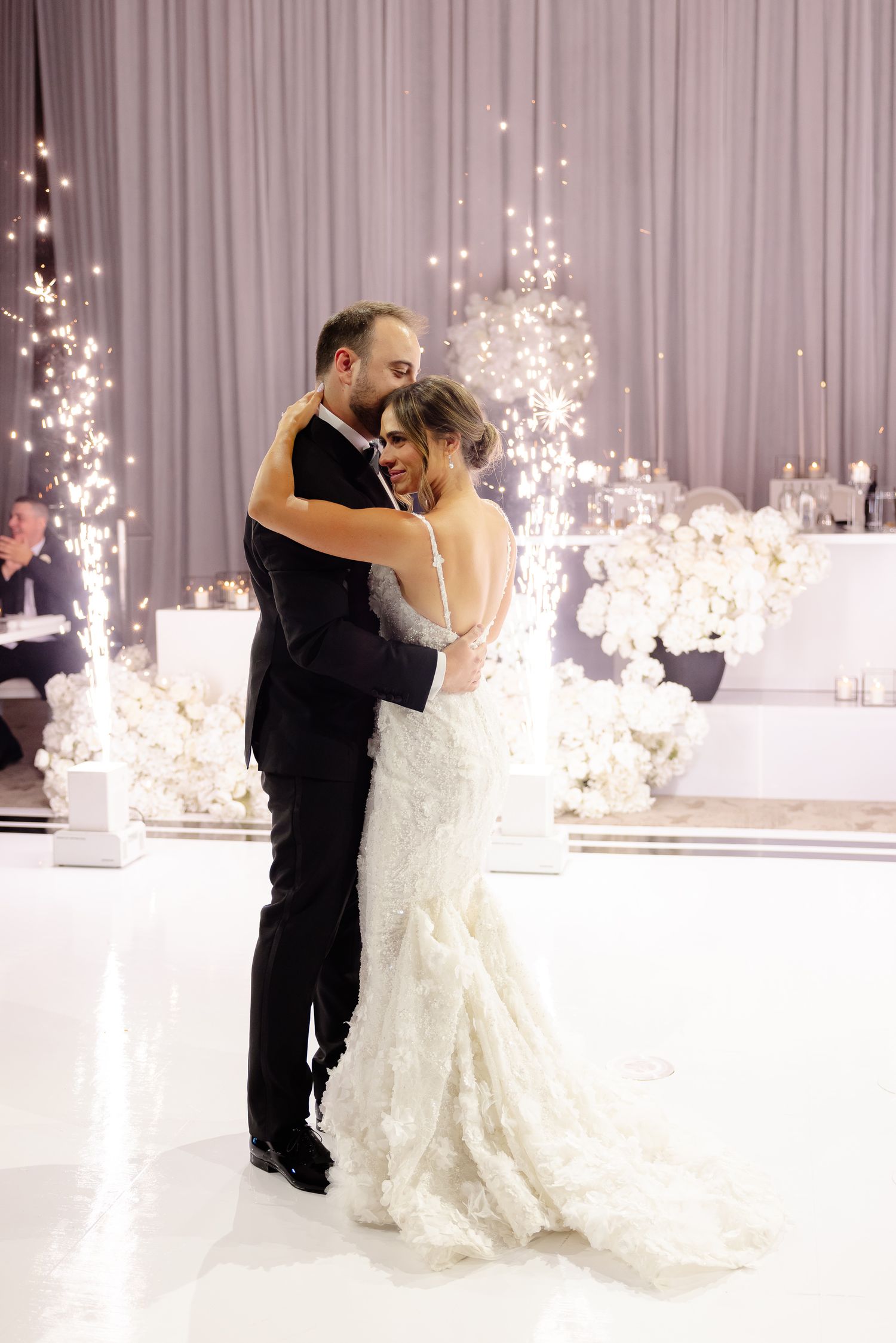 Newly married couple share their first dance in a stunning all-white reception venue decorated with sparkling lights.