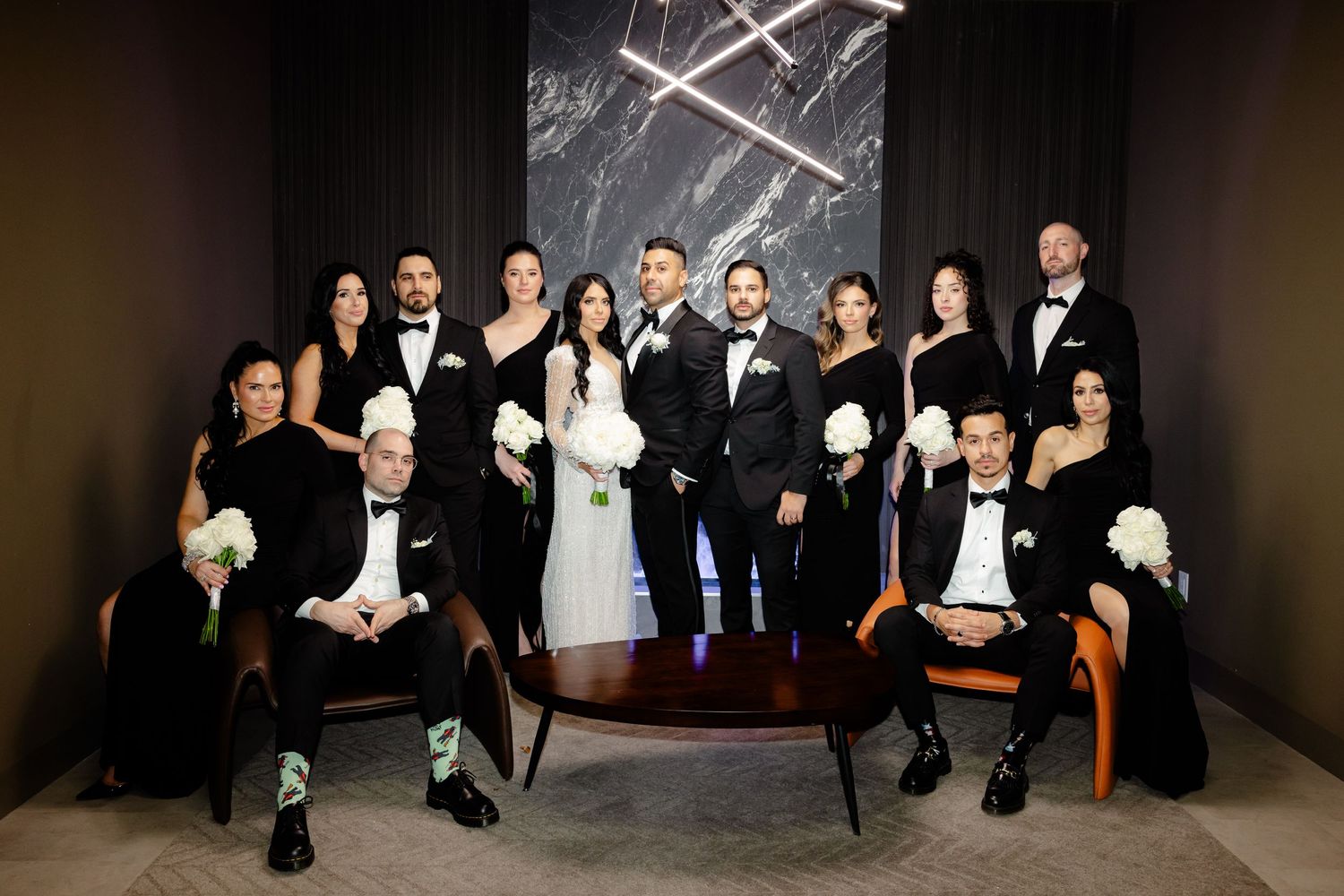 Wedding party poses formally in black attire against dark marble wall with modern lighting fixture.