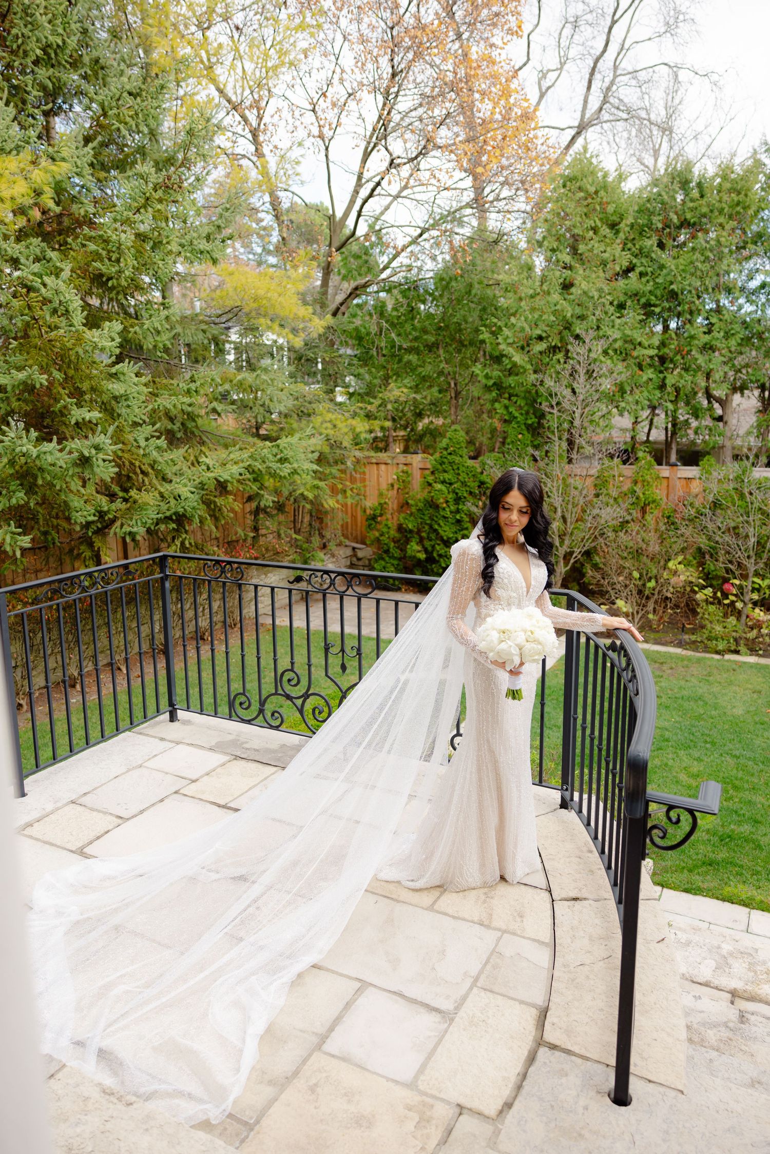 Bride in long-sleeved fitted gown with dramatic cathedral veil poses on stone patio with autumn foliage backdrop.