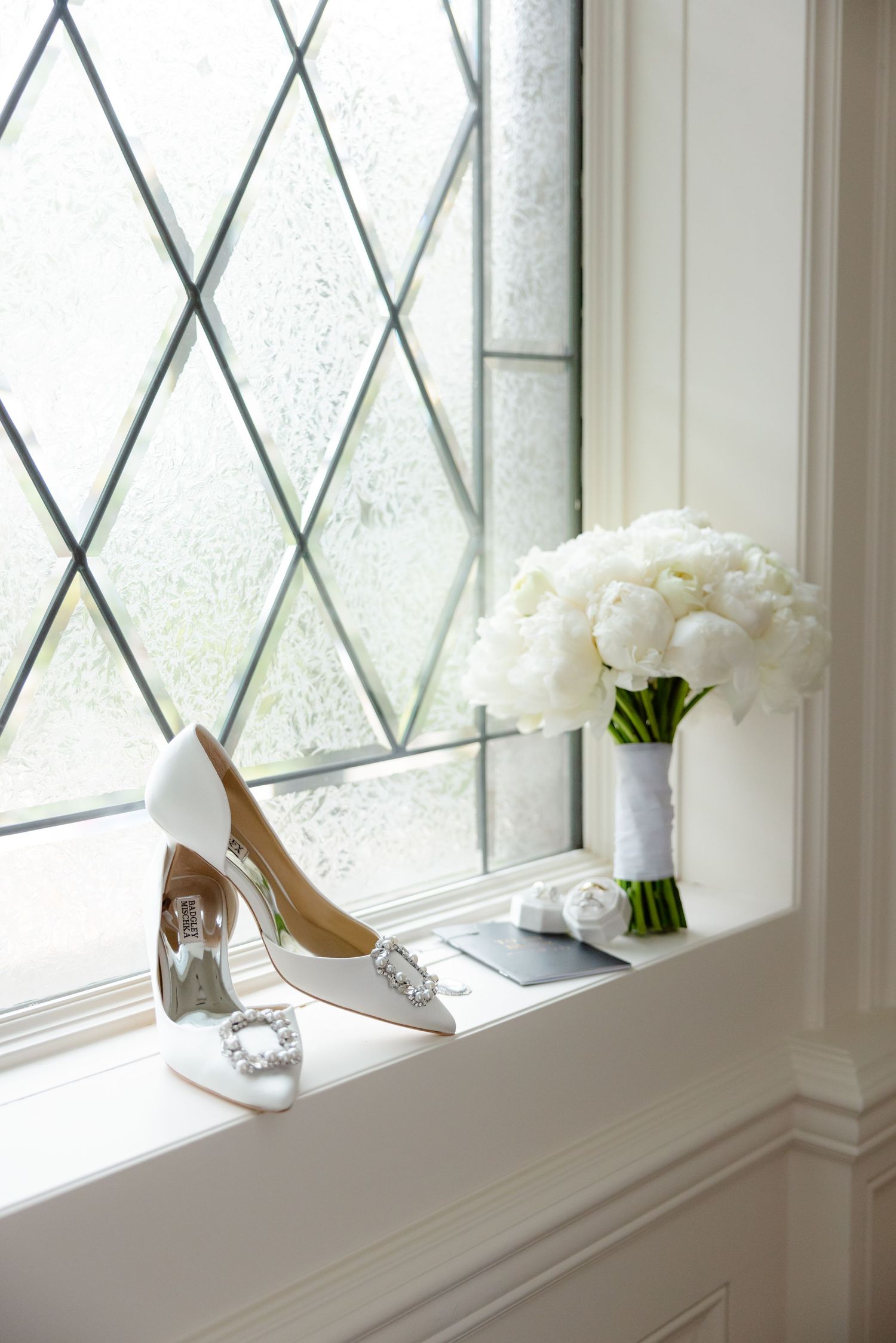 White wedding shoes and bouquet of white peonies on a windowsill with diamond pattern glass.