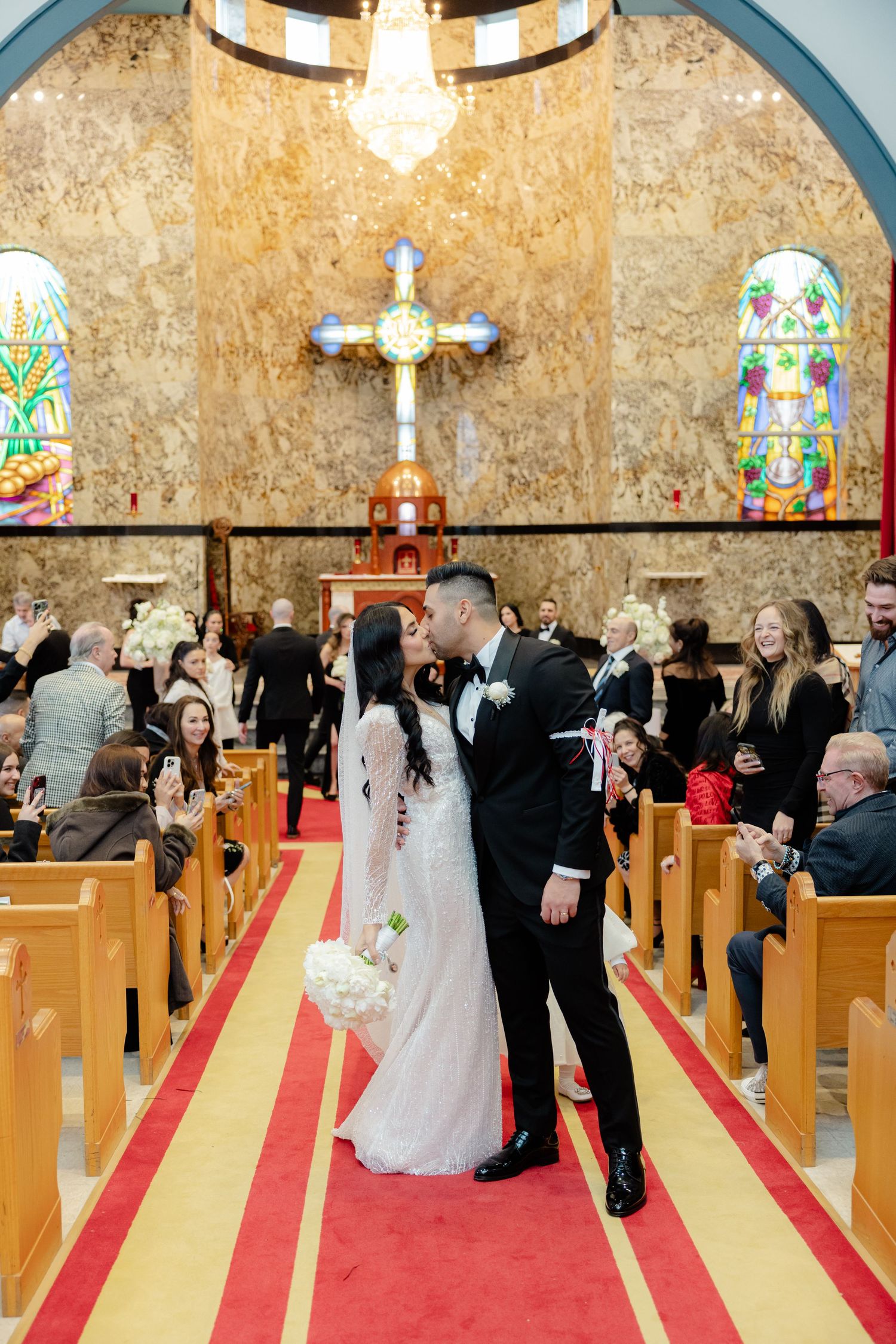 Newlyweds share moment in church aisle with stained glass windows and cross in background.