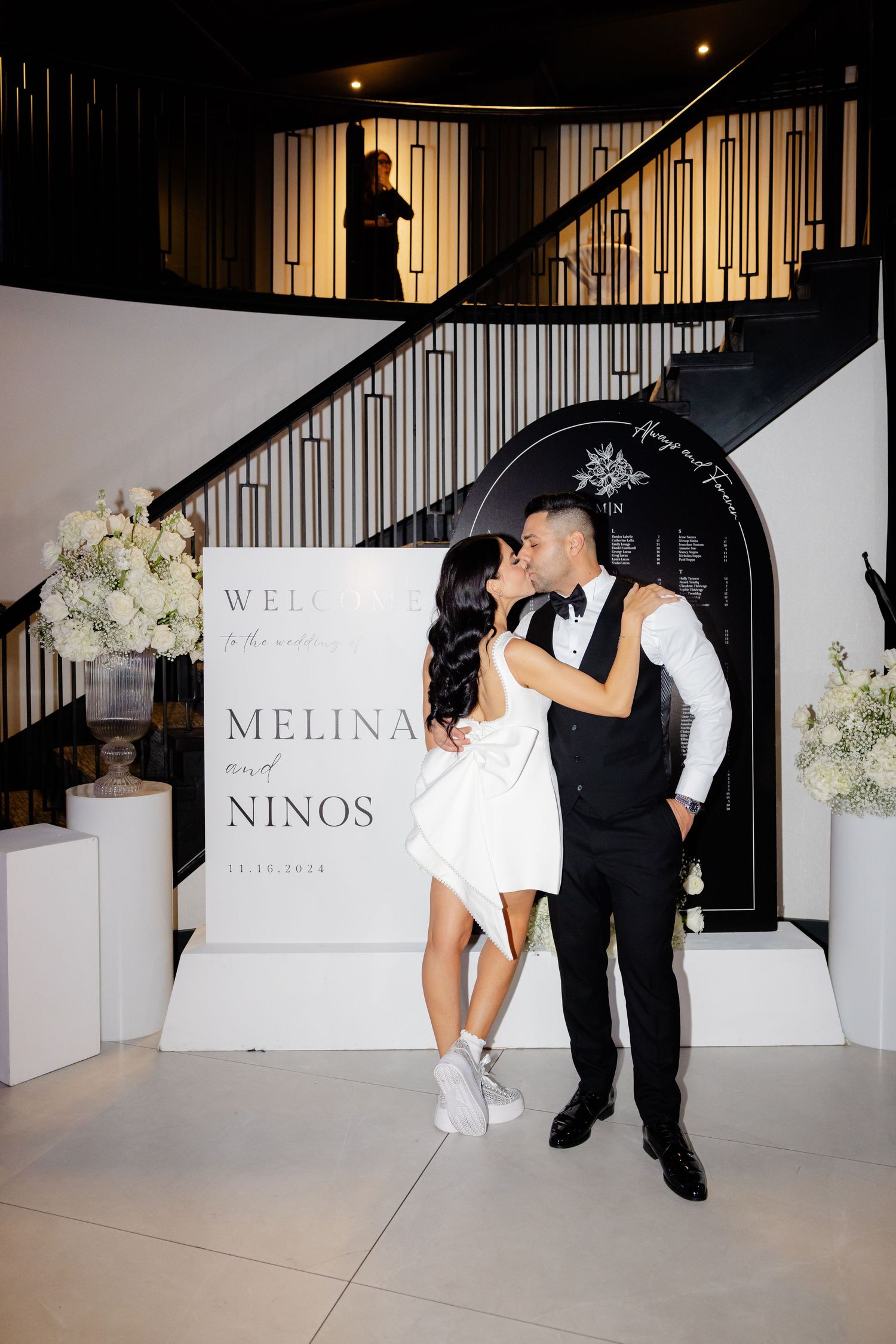 Couple posing at wedding reception in front of elegant signage and floral arrangements.