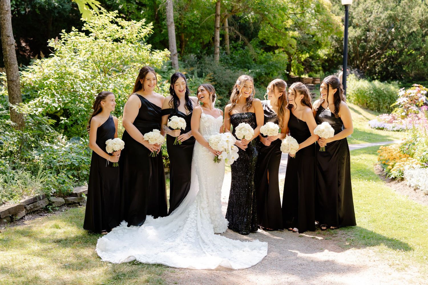 Wedding party poses in beautiful garden setting with bridesmaids in elegant black dresses and holding white bouquets.