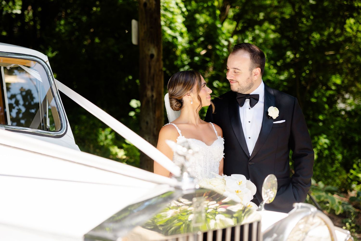 A smiling couple stands beside a vintage Rolls Royce on their wedding day in a lush outdoor setting.