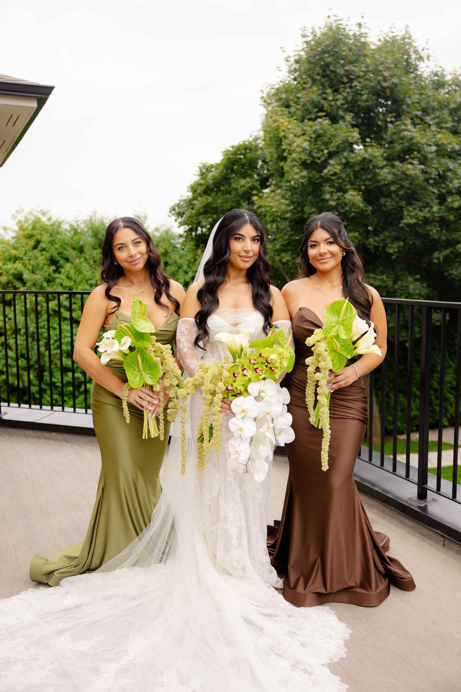 Three bridesmaids in elegant floor-length dresses of olive green and brown pose with bouquets on a balcony.