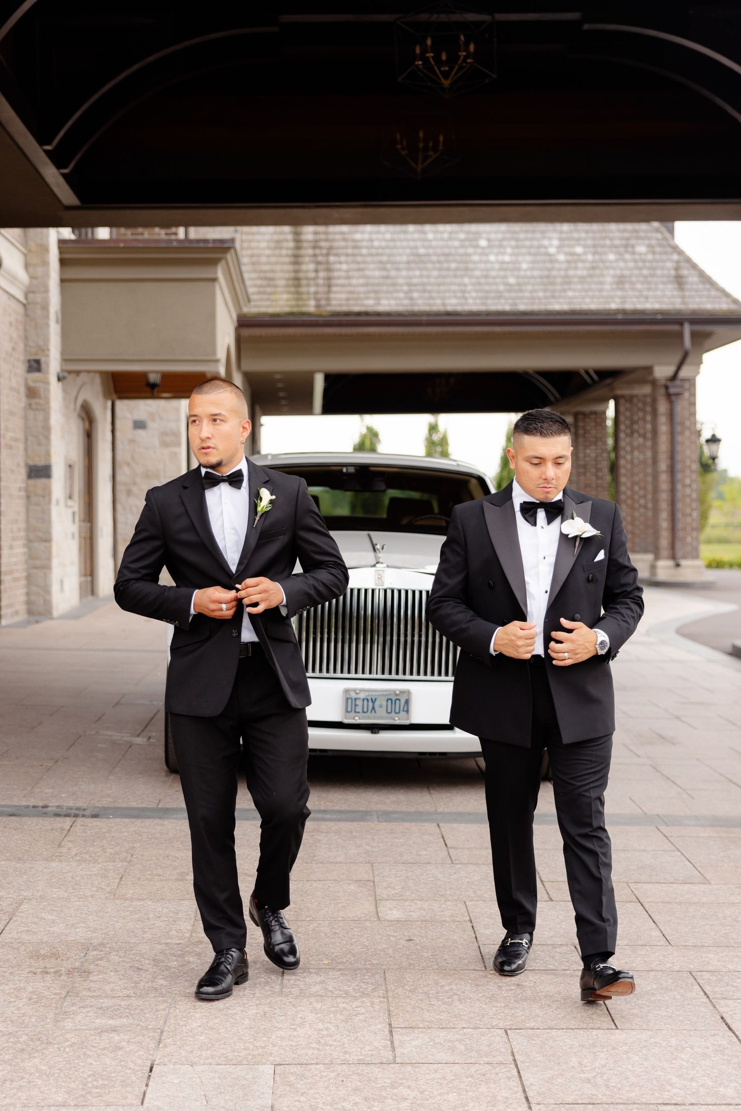 Two groomsmen in black tuxedos and bow ties walk confidently in front of a luxury Rolls Royce at an elegant venue.