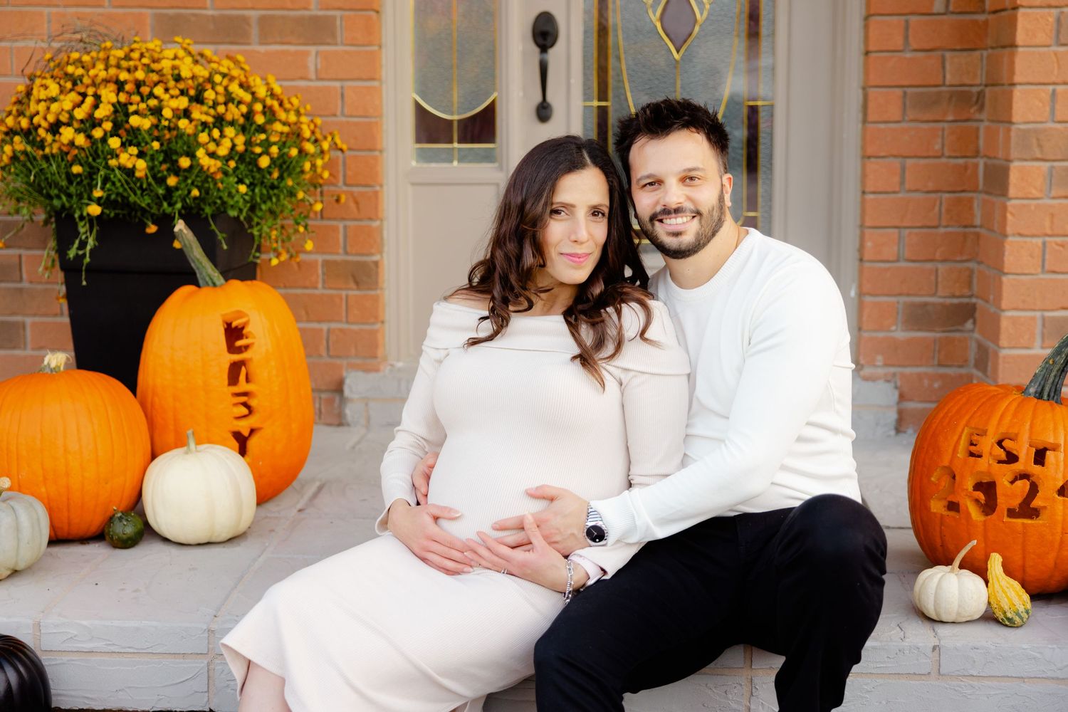 Couple in white sweaters sit on autumn porch with pumpkins and mums, creating a cozy fall scene.