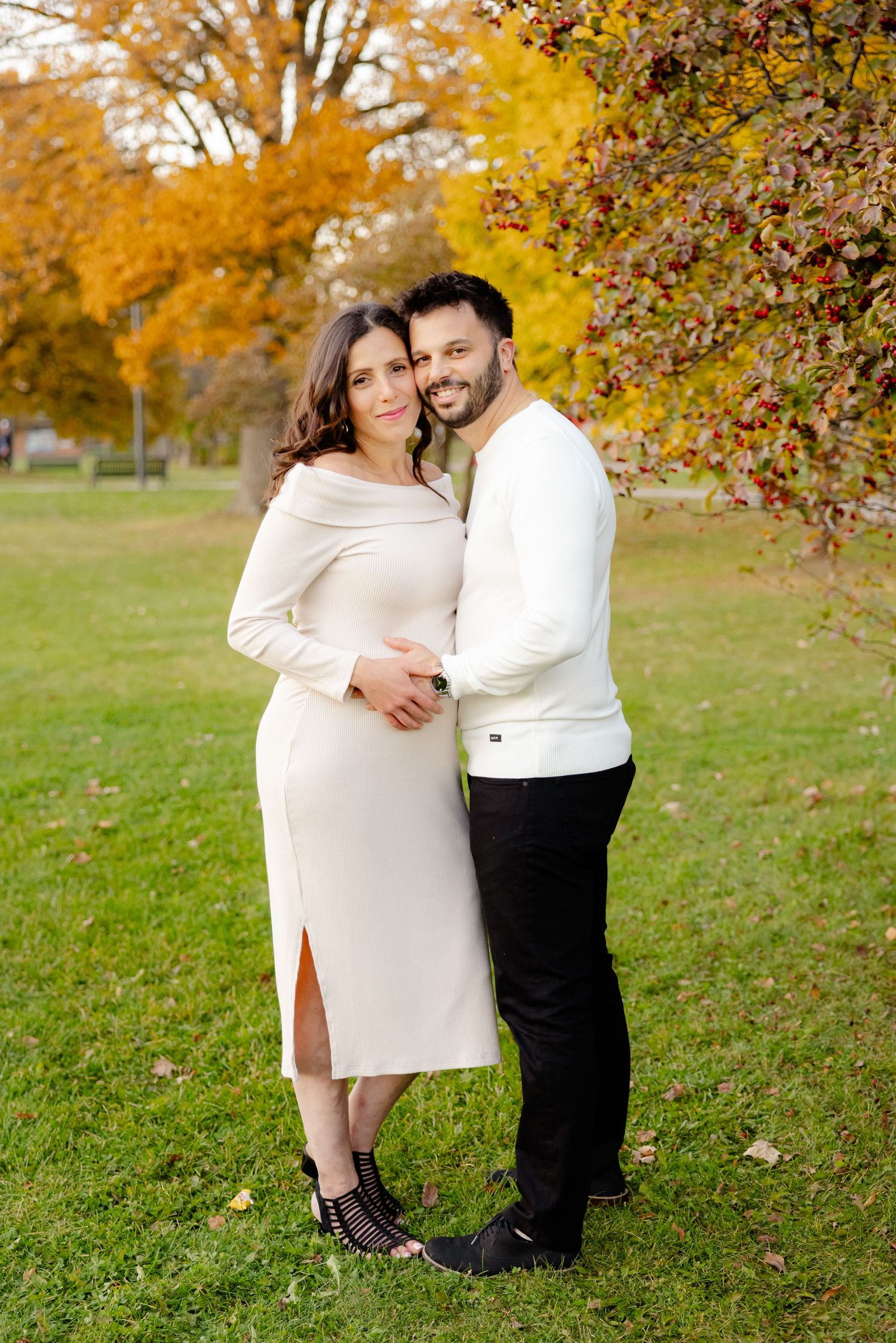 Couple in white outfits pose together on grass with vibrant fall colors in background.