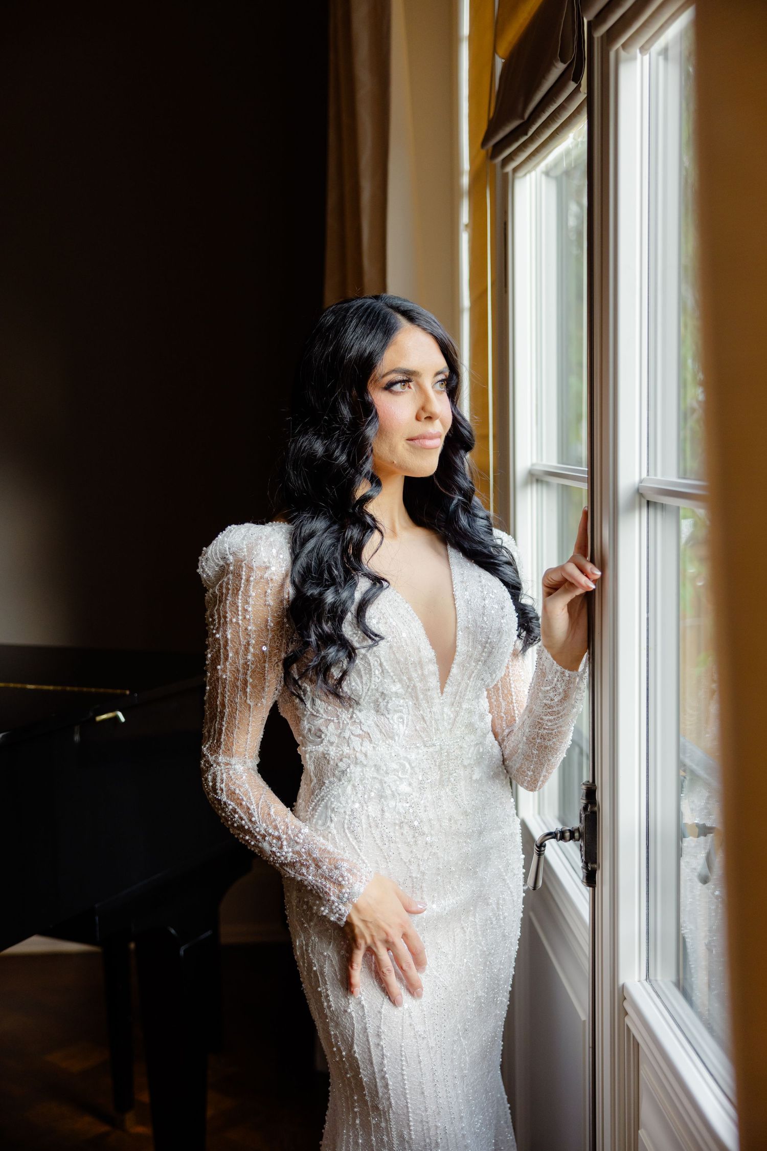 Elegant bridal portrait featuring a form-fitting white beaded gown with long sleeves by a window.