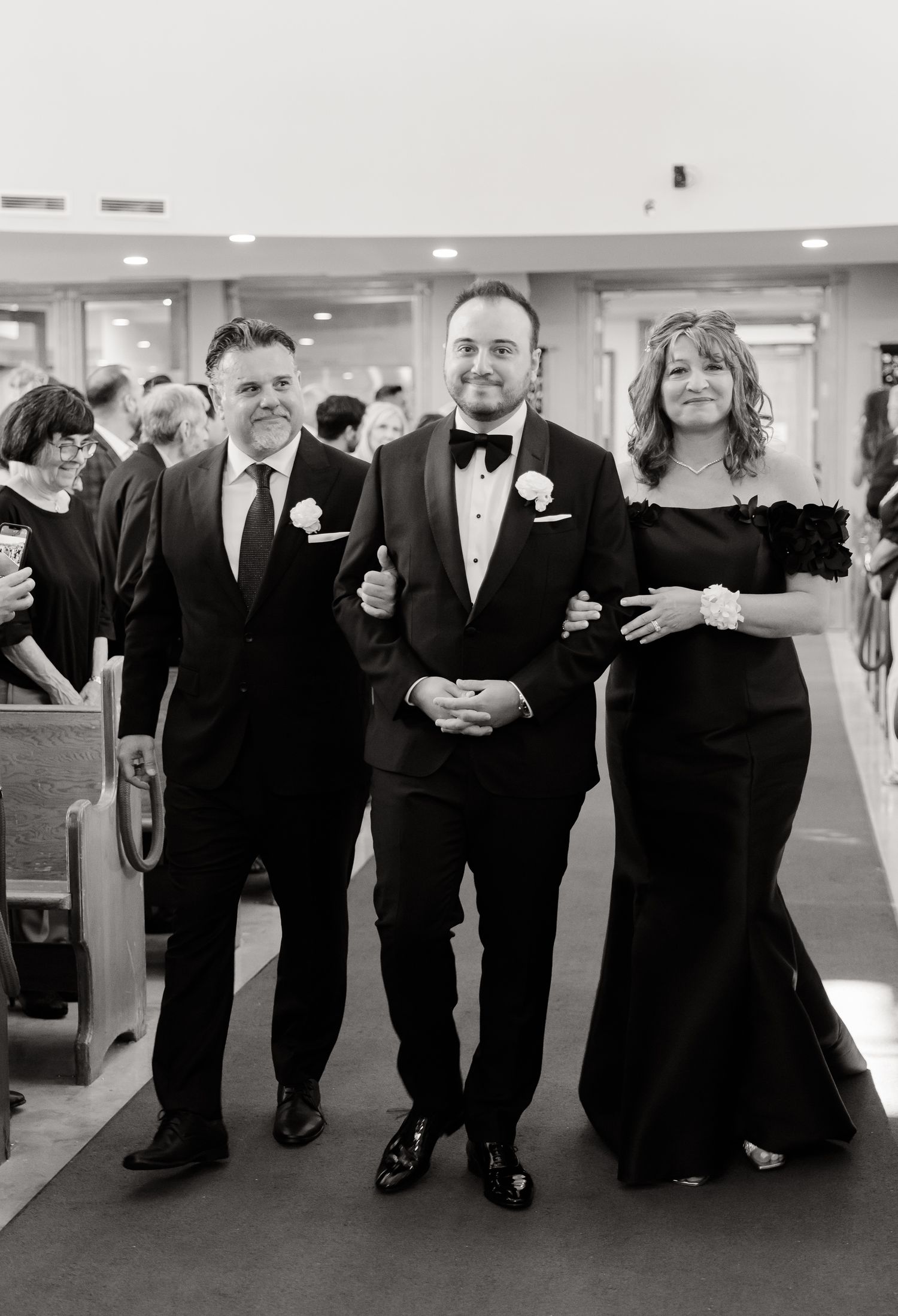 A black and white photograph of three people walking arm-in-arm down a church aisle during a formal event.