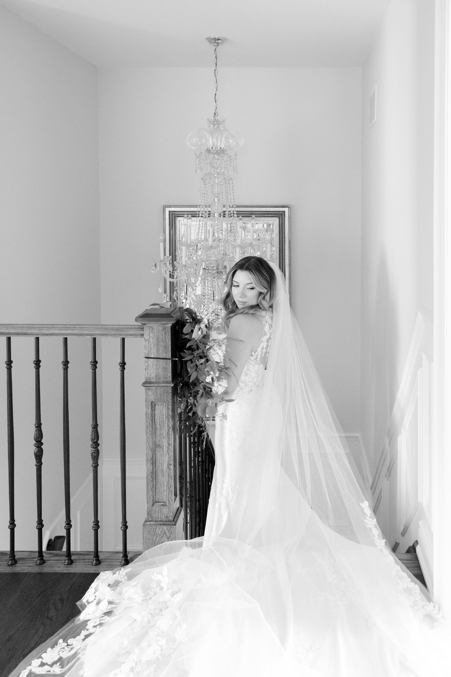 Black and white bridal portrait on staircase with flowing veil and crystal chandelier overhead.