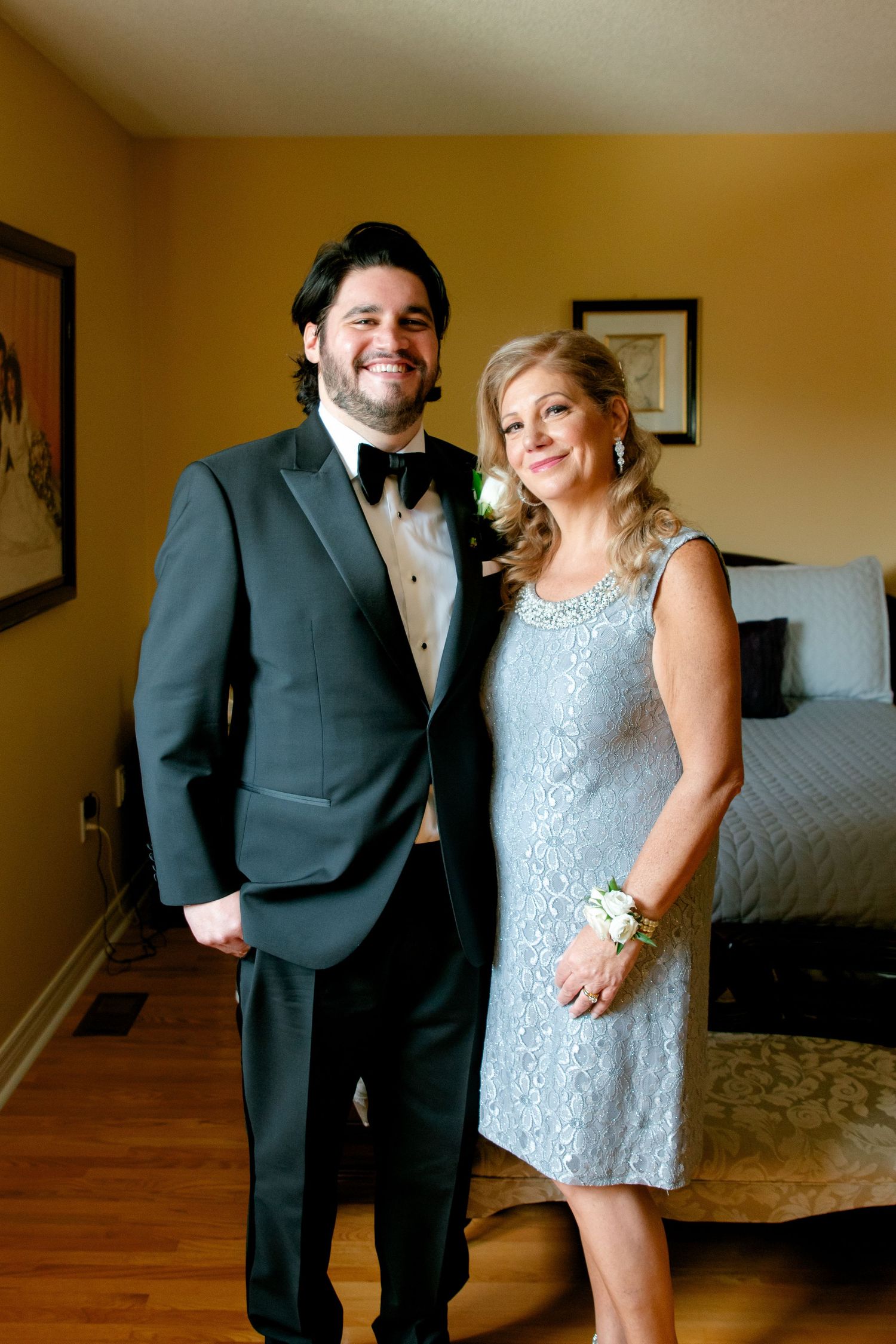 Formal portrait of two people in evening attire standing in front of a bedroom setting.