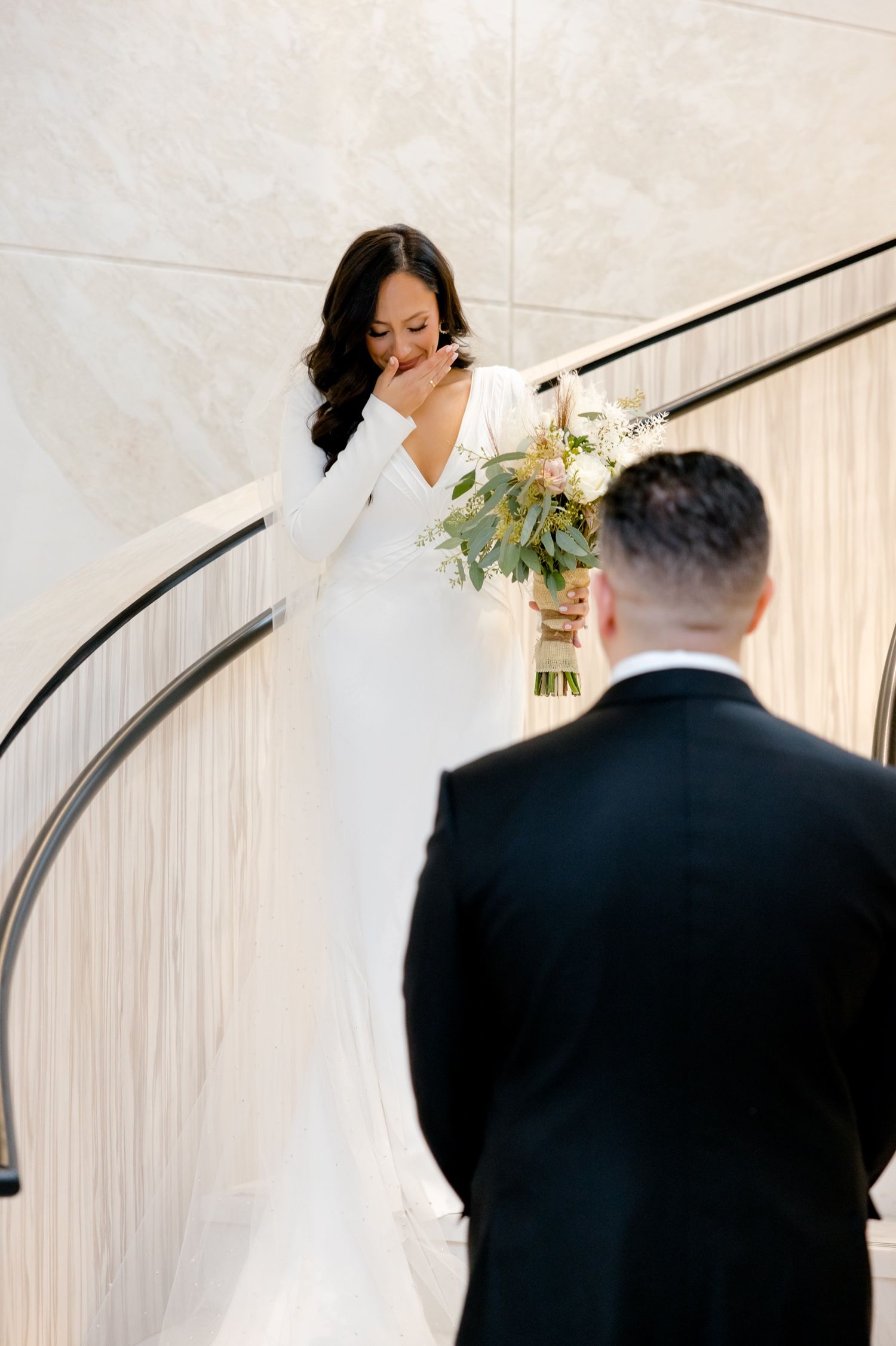 An emotional bride holding a white bouquet during first look on curved staircase.