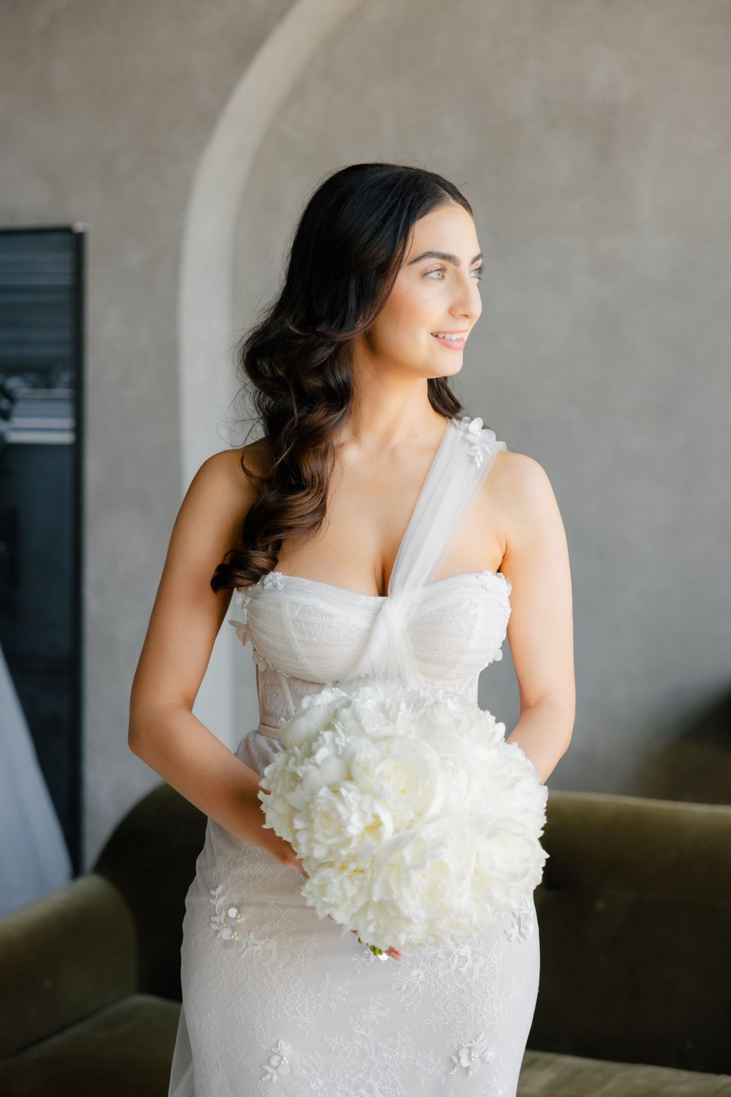 Bride with dark wavy hair holds white bouquet while posing in front of curved wall.